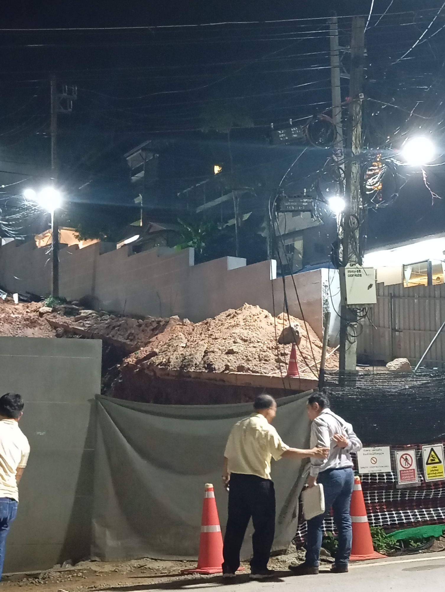 In Karon, Thailand, at night, a construction site is depicted following a landslide. A large pile of dirt and debris obstructs a section of a concrete wall. Two men are engaged in conversation in the foreground. One man, wearing a light yellow shirt and dark pants, gestures towards the other. The second man, in a light blue shirt and blue jeans, holds a white object and appears to be responding. Several orange traffic cones and safety signs are visible, indicating a hazardous area. Utility poles with numerous wires are also present. The scene suggests ongoing work or assessment after the landslide.