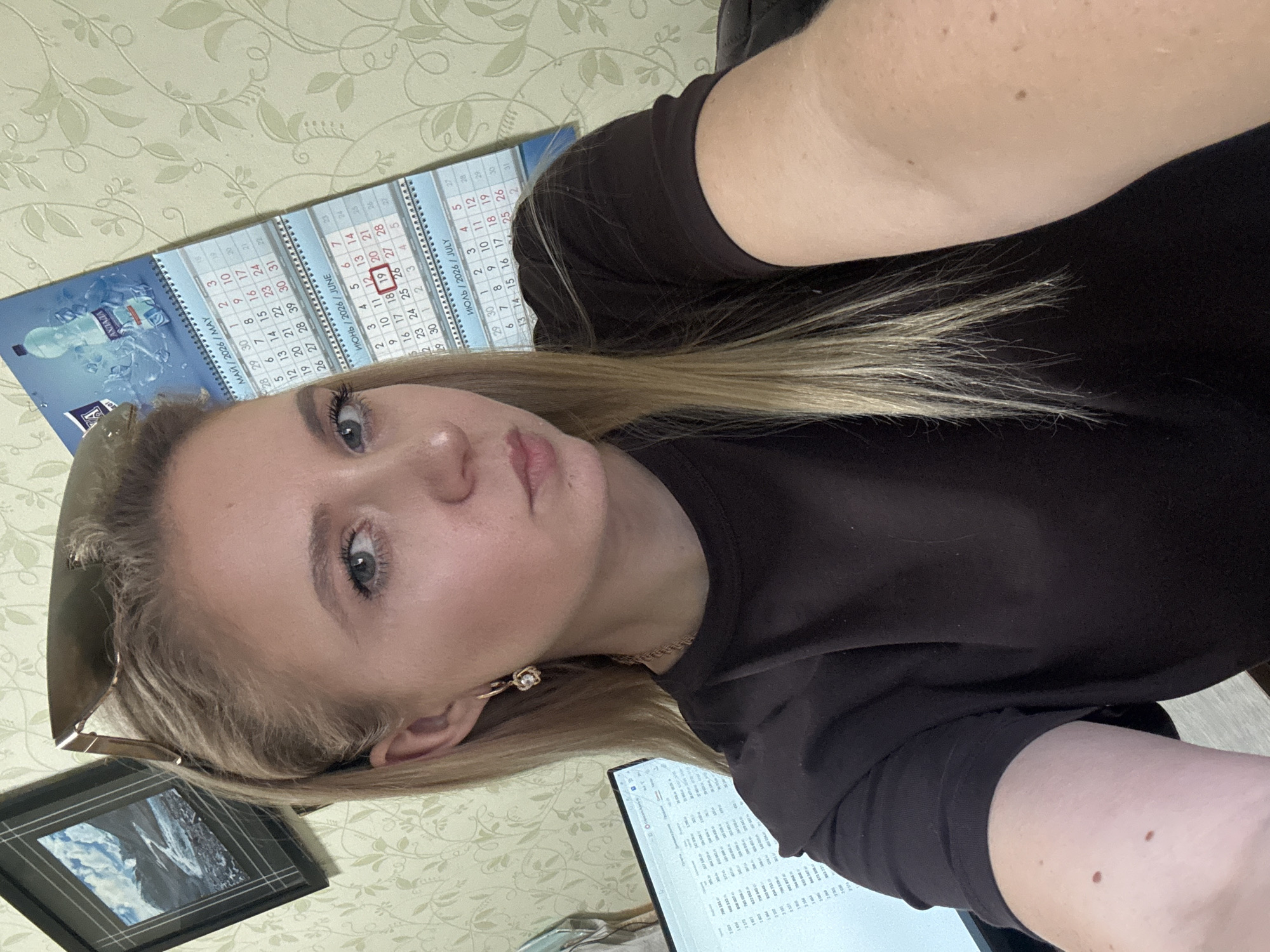 The image shows a young blonde woman with fair skin and blue eyes taking a selfie indoors. She is wearing a dark brown t-shirt and gold earrings. Her sunglasses are resting on top of her head. In the background, a calendar with dates for May, June, and July 2026 is visible, along with a framed picture of mountains and a computer monitor displaying a spreadsheet. The wallpaper in the background has a light green floral pattern. The lighting suggests it is daytime, possibly in an office or home environment.