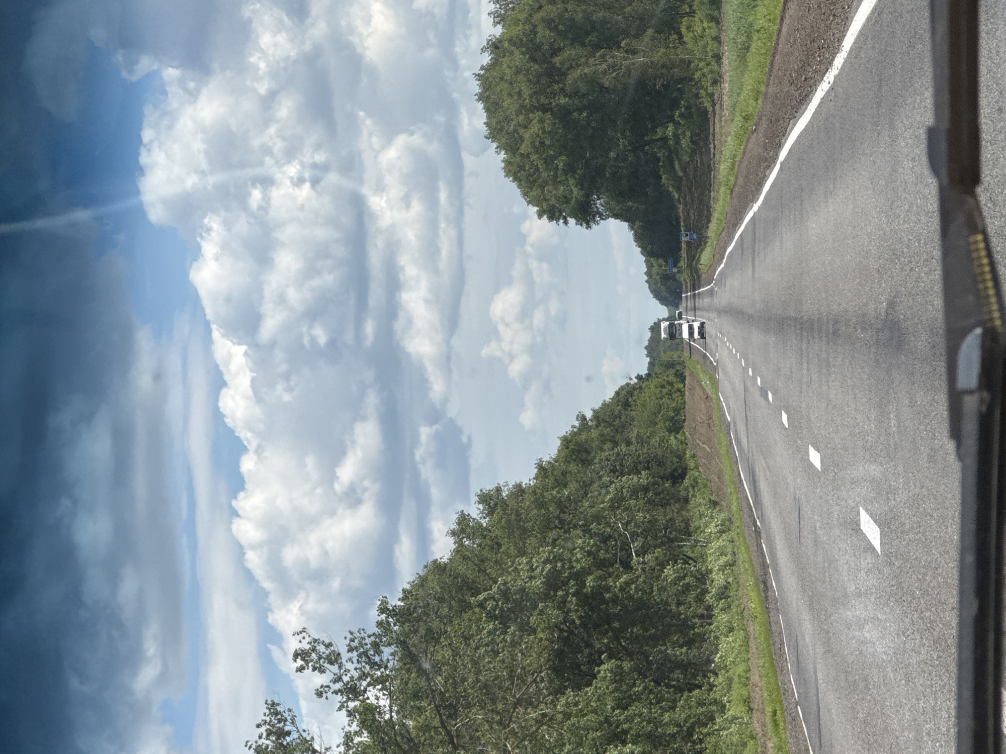 The image depicts a view from a vehicle traveling on a paved road through a rural landscape. The road is marked with white lane dividers. On either side of the road are dense green trees, forming tree lines that extend into the distance. The sky is partly cloudy with large, white cumulus clouds against a blue sky. In the distance, several vehicles are visible on the road, including at least one truck and a white car. The environment appears to be a natural, possibly forested area, characteristic of the countryside. The location is identified as Роўкавіцкі сельскі Савет, Belarus.