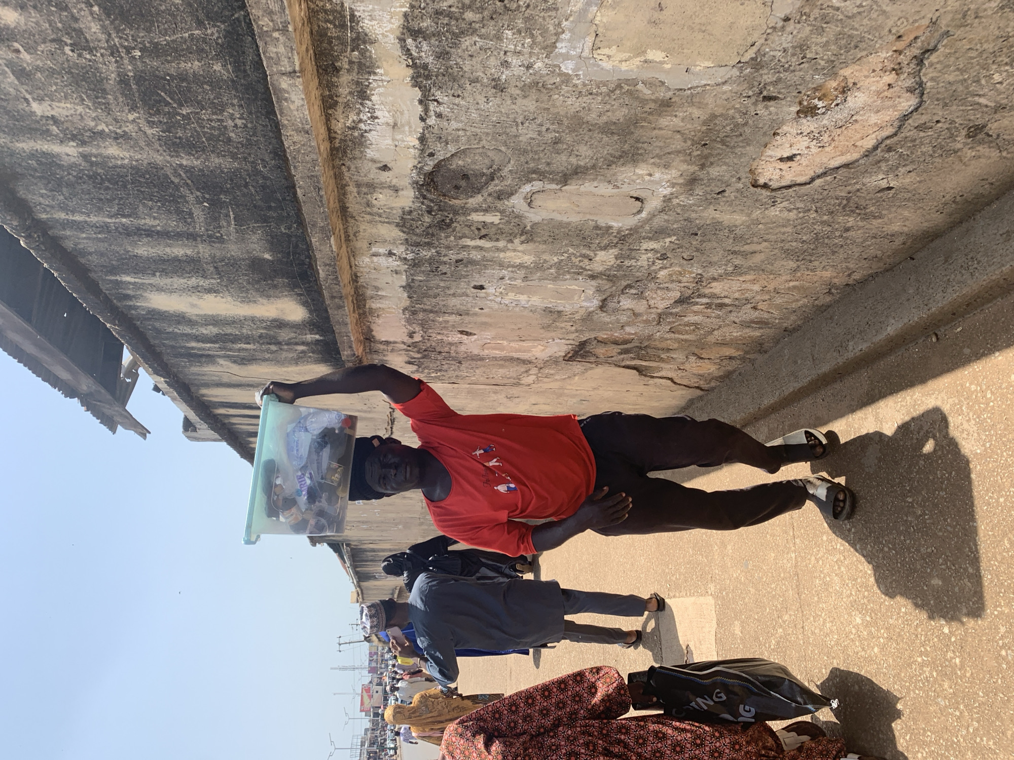 In the city of Jos, Nigeria, a man walks along a dusty sidewalk, carrying a clear plastic tub filled with various items on his head. The tub contains what appear to be plastic bottles and other miscellaneous goods. The man wears a red t-shirt with a graphic design on the front and dark trousers. He is barefoot, wearing sandals. He walks past a weathered, textured concrete wall, which takes up a significant portion of the right side of the frame. Other pedestrians are visible in the background, walking in the same direction along the sidewalk, some wearing traditional clothing and head coverings. The scene is under a bright, sunny sky, casting distinct shadows on the ground. The environment suggests an outdoor market or bustling street scene.