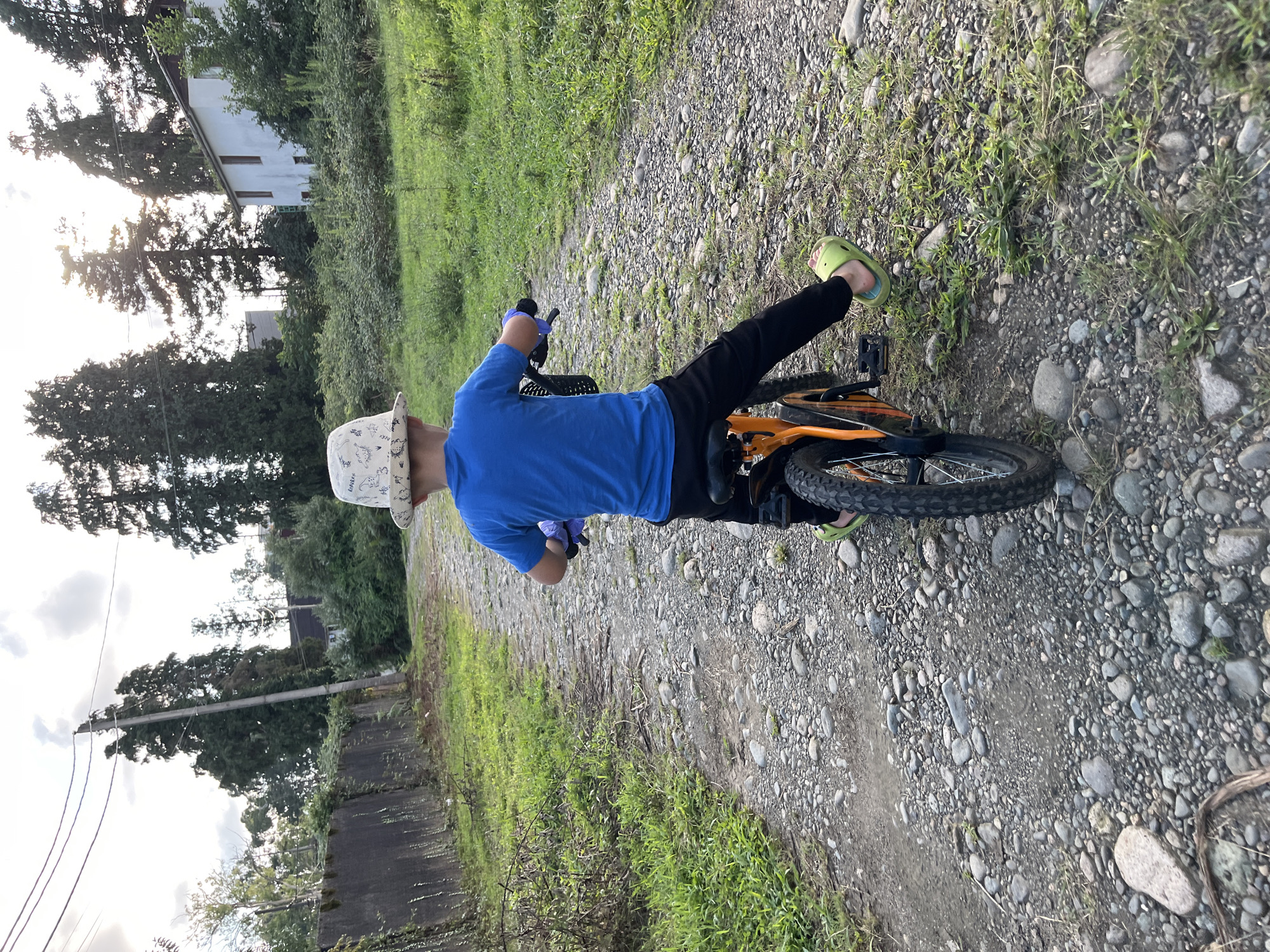 A young boy wearing a blue t-shirt and black pants, and a white hat with a pattern, sits on an orange bicycle on a gravel path in Ureki, Georgia. The boy is facing away from the camera, and his left foot is on the pedal. The path is lined with green grass and trees. In the background, there is a white building and a stone wall. The sky is cloudy.