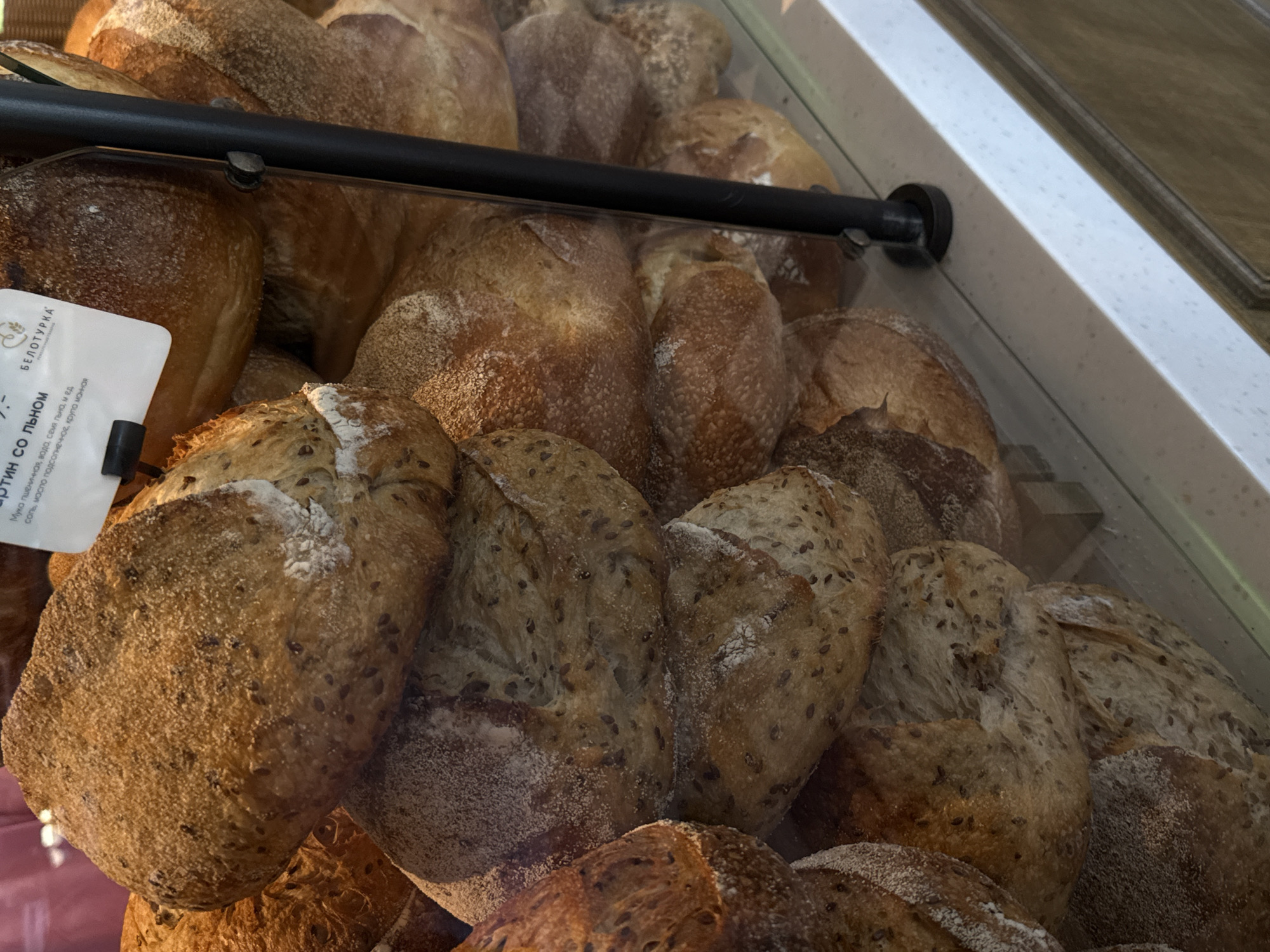 A display case filled with various types of bread is shown in a medium, eye-level shot. The bread is primarily sourdough with flax seeds. A small white label reading "БЕЛОТУРКА" and "Львом" is attached to one of the loaves. A black bar runs horizontally across the middle of the display case. The scene is likely located in Izhevsk, Russia.