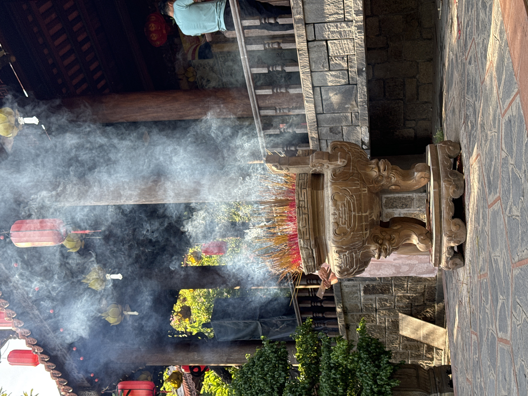 The image depicts an outdoor scene at the Long Son Pagoda in Nha Trang, Vietnam. In the foreground, a large, ornate incense burner sits on a stone base, filled with numerous burning incense sticks. Thick white smoke billows upwards from the incense, obscuring parts of the background. Behind the incense burner, a stone wall and a wooden railing are visible. A person in a light blue shirt and dark pants is standing on a raised wooden platform to the right, with their back to the camera. Several red lanterns are hanging from the eaves of the pagoda, some of which are partially obscured by the smoke. Green trees and foliage can be seen in the background, along with architectural elements of the pagoda, including wooden pillars and a tiled roof. The ground is paved with stone tiles. The overall impression is one of a sacred space with active devotional practices.