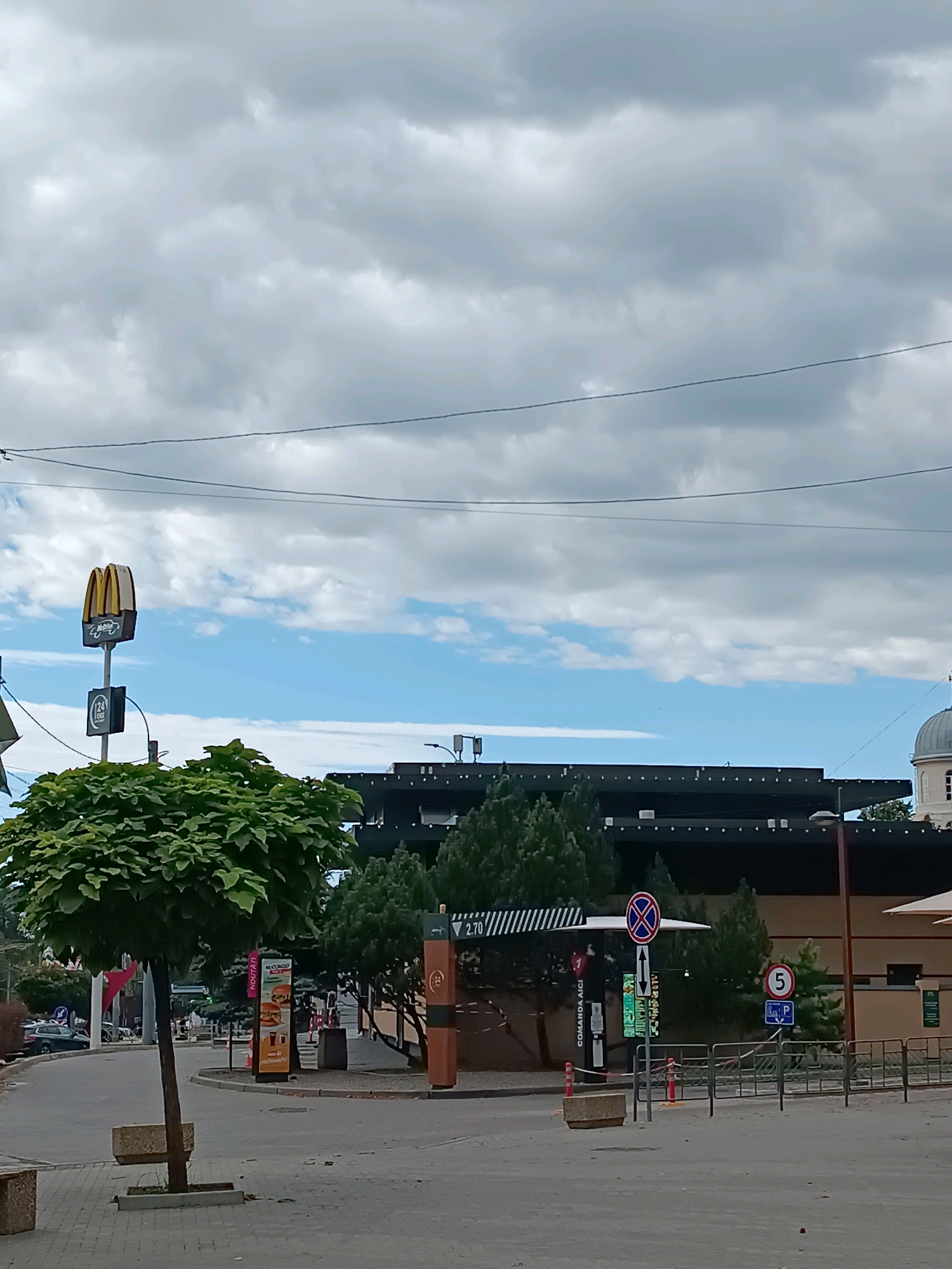 The image shows the exterior of a McDonald's restaurant, likely a drive-thru, in Chișinău, Moldova. The McDonald's sign with the golden arches is visible on a tall pole on the left side of the image, above a green tree. The building has a dark facade with a white dome structure visible in the upper right corner, suggesting proximity to a church or similar building. Several traffic signs are present, including a no-parking sign with arrows and a speed limit sign with the number '5'. Menu boards and drive-thru ordering stations are also visible. The area appears to be a paved public space with some greenery, including trees and planters. The sky is partly cloudy. There are no people clearly visible in the image.