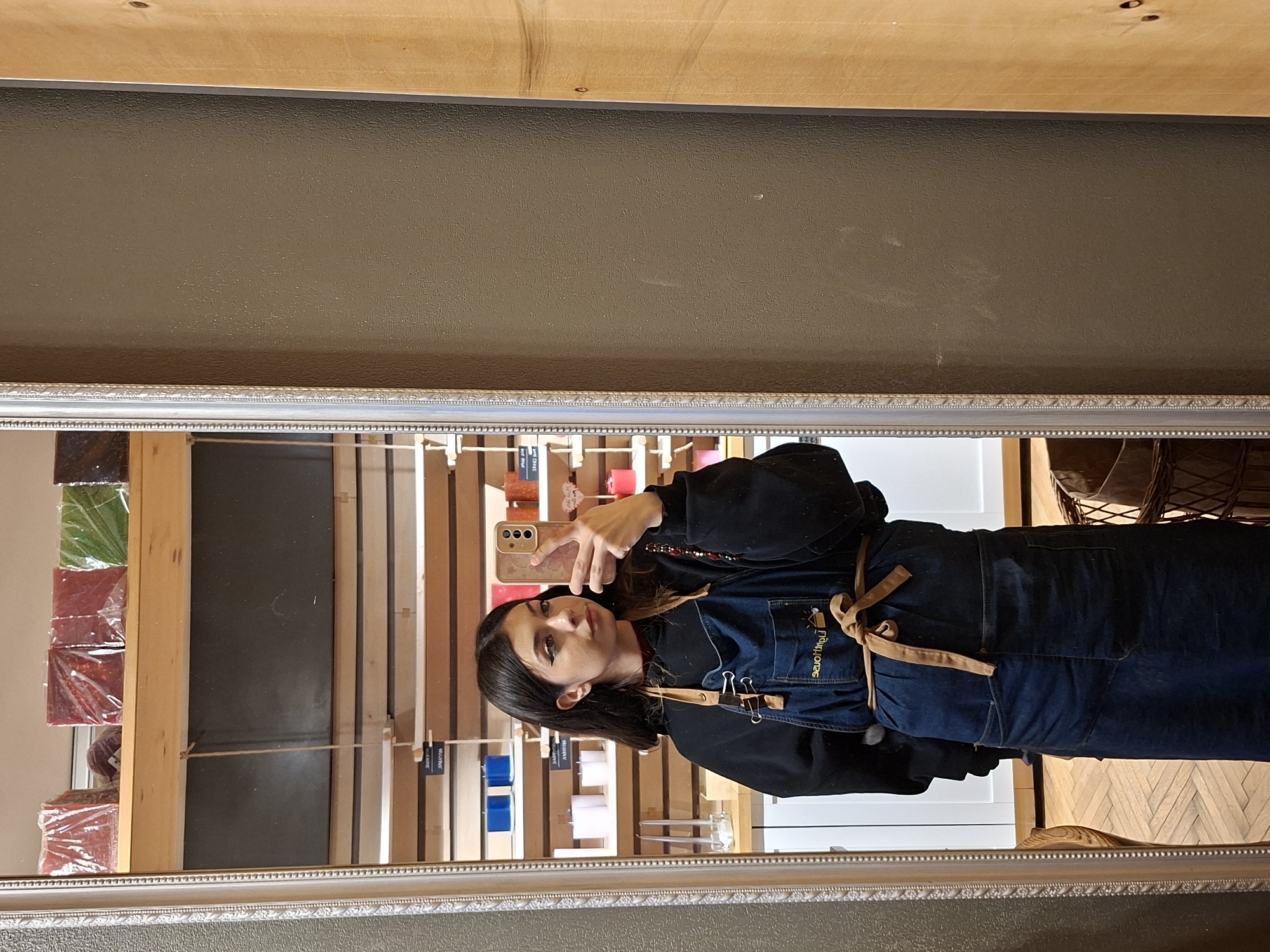The original image is oriented horizontally, depicting a mirror selfie of a young woman in what appears to be a retail or craft shop. If rotated upright, the woman stands in the reflection of a large mirror, framed with an ornate, silver-colored border.
The woman has dark, shoulder-length hair and is wearing a dark, long-sleeved top underneath a dark blue denim apron. The apron features light brown straps tied at the waist and a yellow logo on the chest that reads "Play Jones" with a small graphic. She holds a light-colored smartphone with a patterned case up to take the photo, partially obscuring her face but revealing her dark eyeliner and a composed expression with a slight smile.
The background visible in the mirror's reflection showcases an indoor setting. On the left side of the reflection, stacks of rectangular items, likely candles or soaps, are wrapped in clear plastic and are visible in colors such as red, green, and dark red/purple. Further into the background, wooden shelves display numerous small items, including white, blue, and pink blocks or containers. Small labels hang from the shelves, with some faint text suggesting words like "SQUARE" and "ROUND" on specific items. The flooring reflected in the lower right is a light-colored wood laid in a herringbone pattern. Above the mirror frame, a dark, textured wall is visible, with a wooden ceiling or upper wall panel above it.
The setting suggests a commercial space, possibly an artisan shop or gift store in Sofia, Bulgaria. The apron indicates the woman may be an employee or artisan.