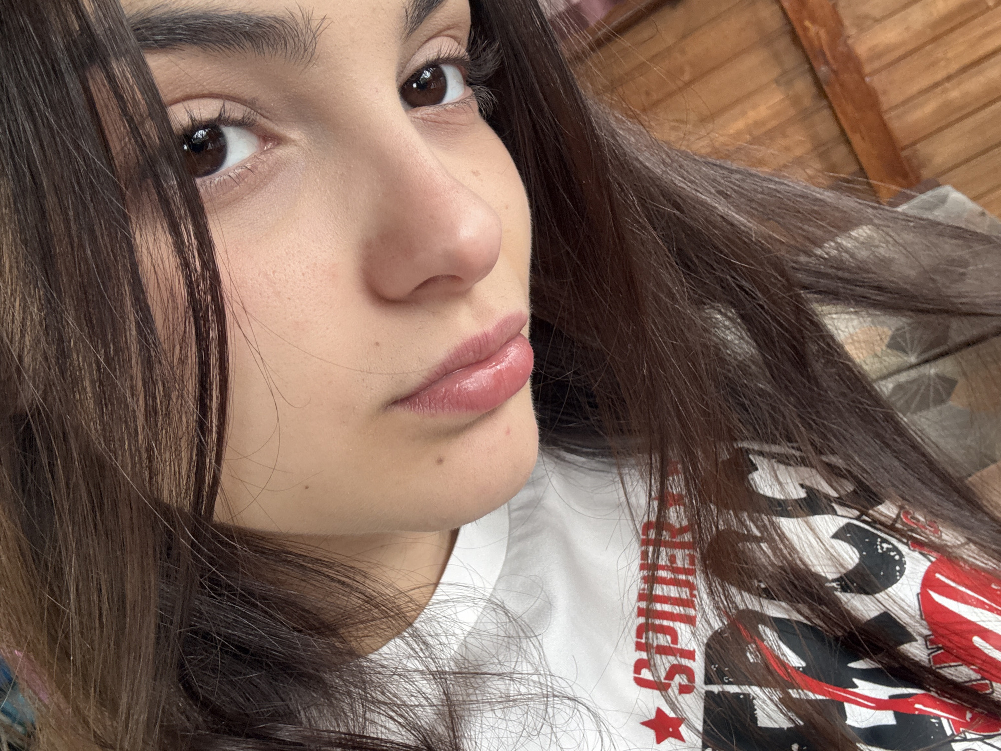 A close-up, angled portrait of a young woman with dark brown hair and brown eyes. She is facing slightly to the left of the frame. The background is blurred, but there is a wood ceiling visible in the upper right, a pattern of pillows or cushions, and a white t-shirt with the word "Spider" written on it in red. There is no clear indication of the location being in Polena, Bulgaria.