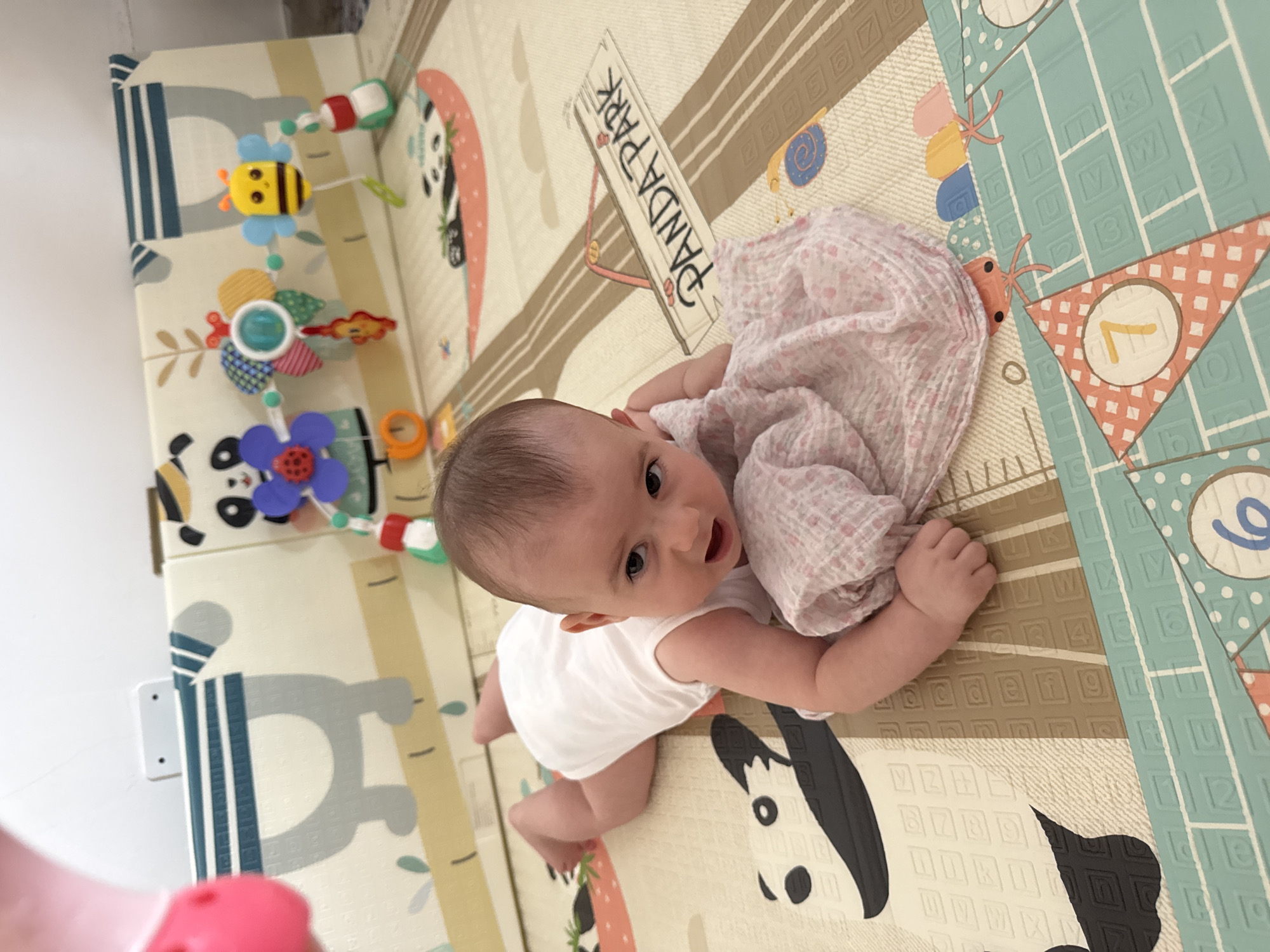 A baby is on its stomach on a play mat. The baby is wearing a white onesie. The play mat has pandas and other cartoon characters. The baby has a pink and white blanket near its head. The background has toys attached to a mat. The scene appears to be indoors.