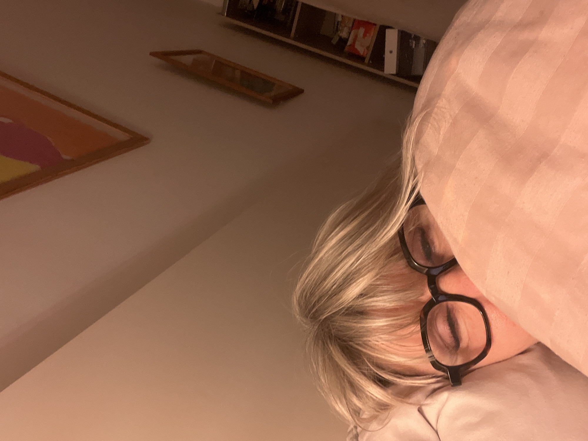 The image shows a person lying in bed with their face partially obscured by a pillow. The person has blonde hair and is wearing black-rimmed glasses. The person is lying on a white pillow with striped pink bedsheets. There are framed pictures hanging on the wall, and a shelf can be seen above the person's head. The location in Chambœuf, France, is not verifiable from the image.