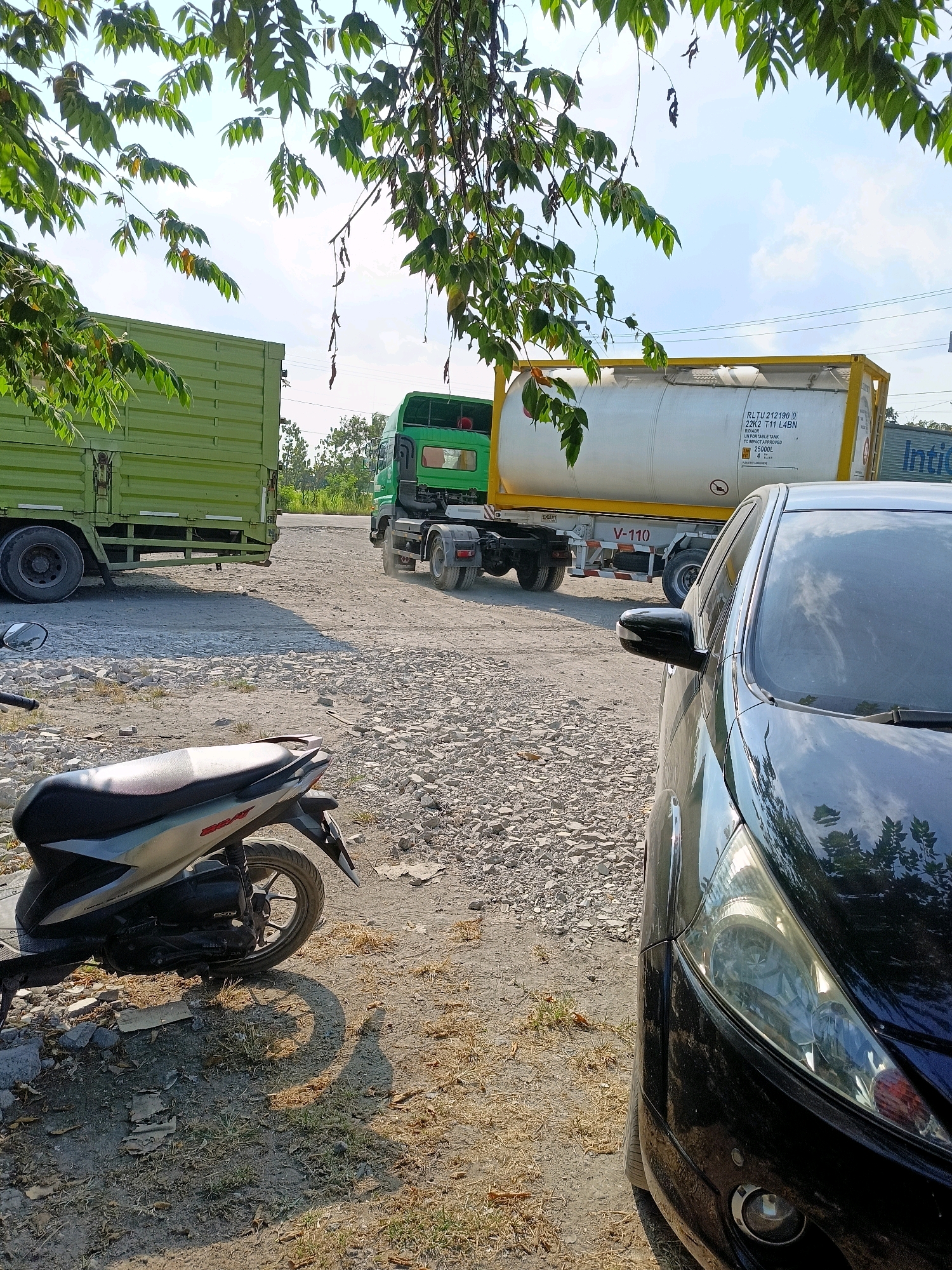 The image depicts a dry, unpaved lot with several vehicles. In the foreground on the left, a silver and black scooter is parked. To the right, the front fender and headlight of a black car are visible. In the middle ground, a large green truck is parked on the left, and a white tank truck with a yellow frame is on the right. The tank truck has markings indicating "RLTU 212190 0 22K2 T11 L4BN" and "UN PORTABLE TANK TC-IMPACT APPROVED 25000L". Overhead, green leaves from a tree partially obscure the bright blue sky. The environment appears to be an outdoor industrial or service area in Kemudo, Indonesia, characterized by gravel and dirt ground.
