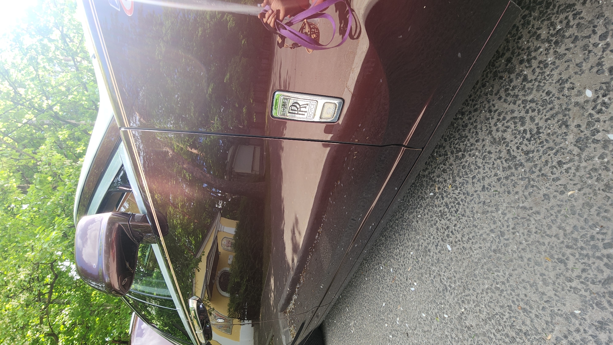 The image depicts a portion of a maroon Rolls Royce parked on a gray asphalt surface, likely a street. The car's emblem is visible on the side panel. Reflections on the car's surface show trees and a building. A purple strap is partially visible on the car's door. It is possible that the image was taken in Khimki, Russia, based on contextual information or location data associated with the image.