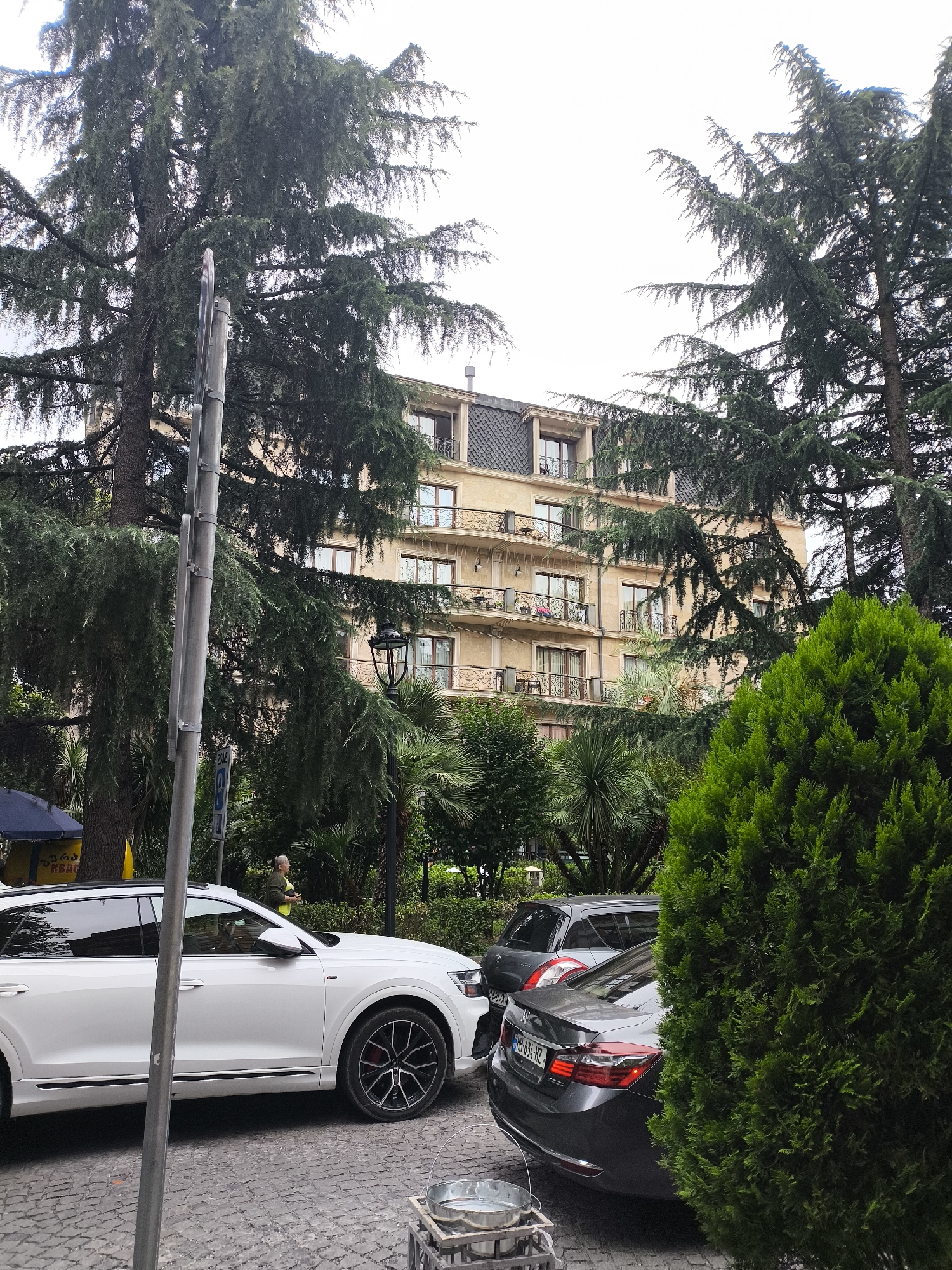 The image depicts a street scene in Batumi, Georgia. In the foreground, a white Audi SUV is parked on a cobblestone street. To its right, a dark-colored sedan is visible, partially obscured by a large, manicured green bush. Behind the vehicles, a multi-story building with beige walls and dark-tiled roofs is seen, featuring balconies with ornate railings. Tall evergreen trees with dense foliage frame the building and the street. A lamppost stands near the center of the frame, and a person in a yellow vest is visible in the background, standing near a blue awning. The sky is overcast.
