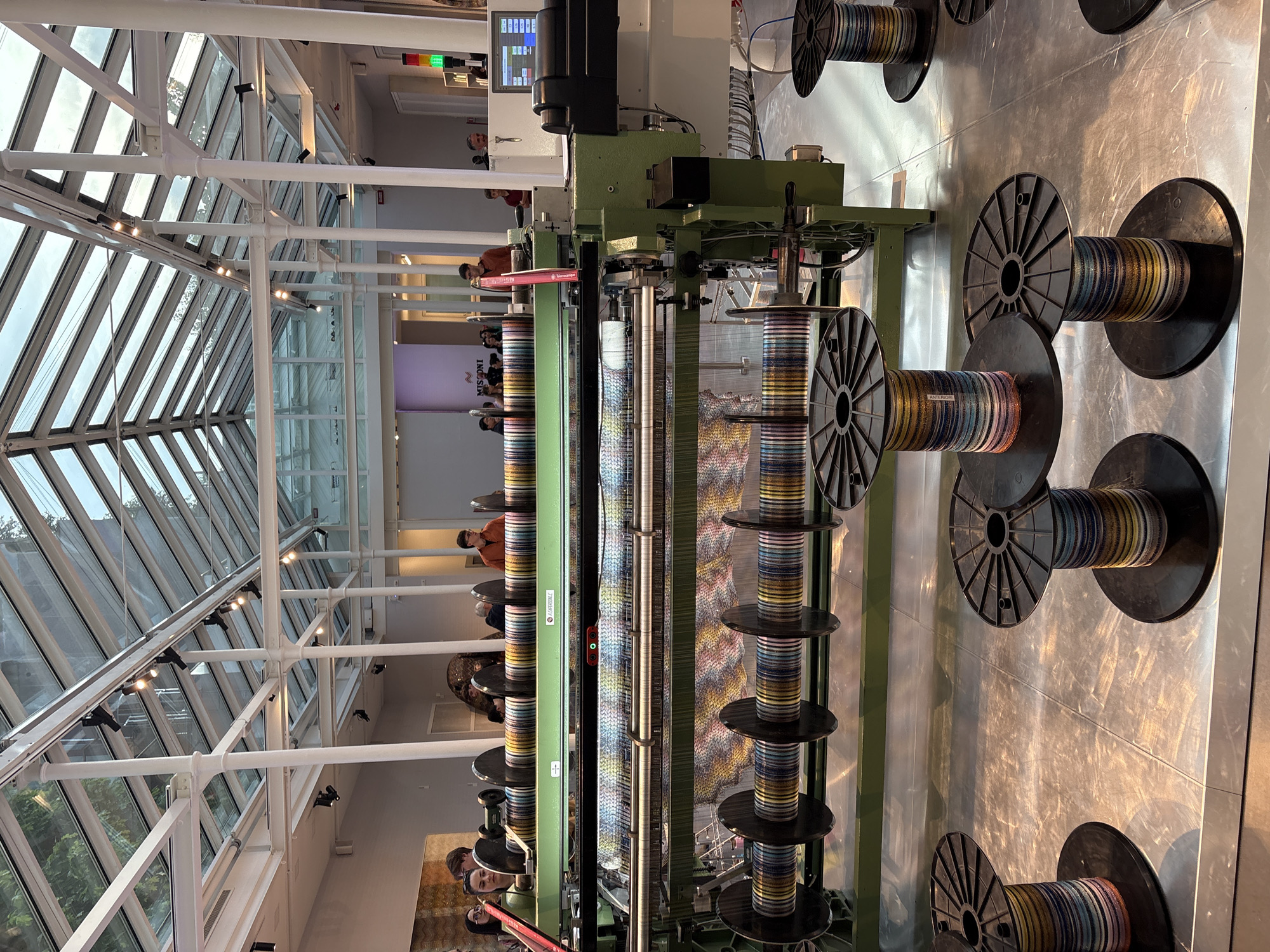 The image shows a weaving machine on display at the Fondazione Missoni museum in Milan, Italy. The machine is predominantly green and black, with numerous spools of colorful thread arranged along its structure. The threads are fed into a loom creating a woven textile with colorful stripes and patterns. People are observing the machine in the background. The museum has a large, high ceiling with exposed white structural supports and glass windows.