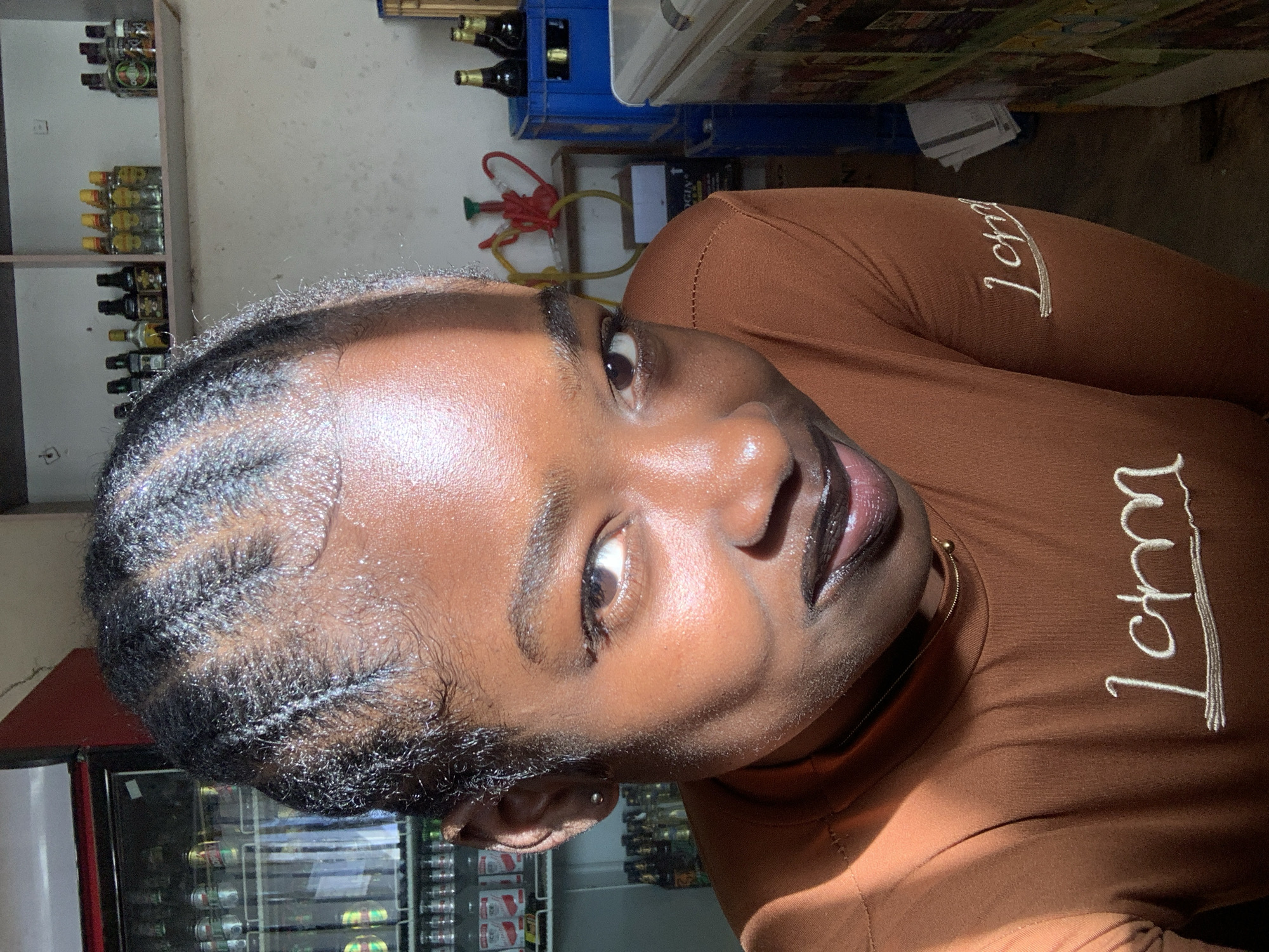 A young woman with braided hair is visible in a partial selfie. She is wearing a brown long-sleeve shirt with the word "Ichi" embroidered on it. The setting is indoors, possibly a store, as shelves stocked with bottles and a refrigerated display case filled with beverages are visible in the background. The location is identified as New Stadium, Nigeria. The lighting is inconsistent, with the top of her head in shadow and her face illuminated.