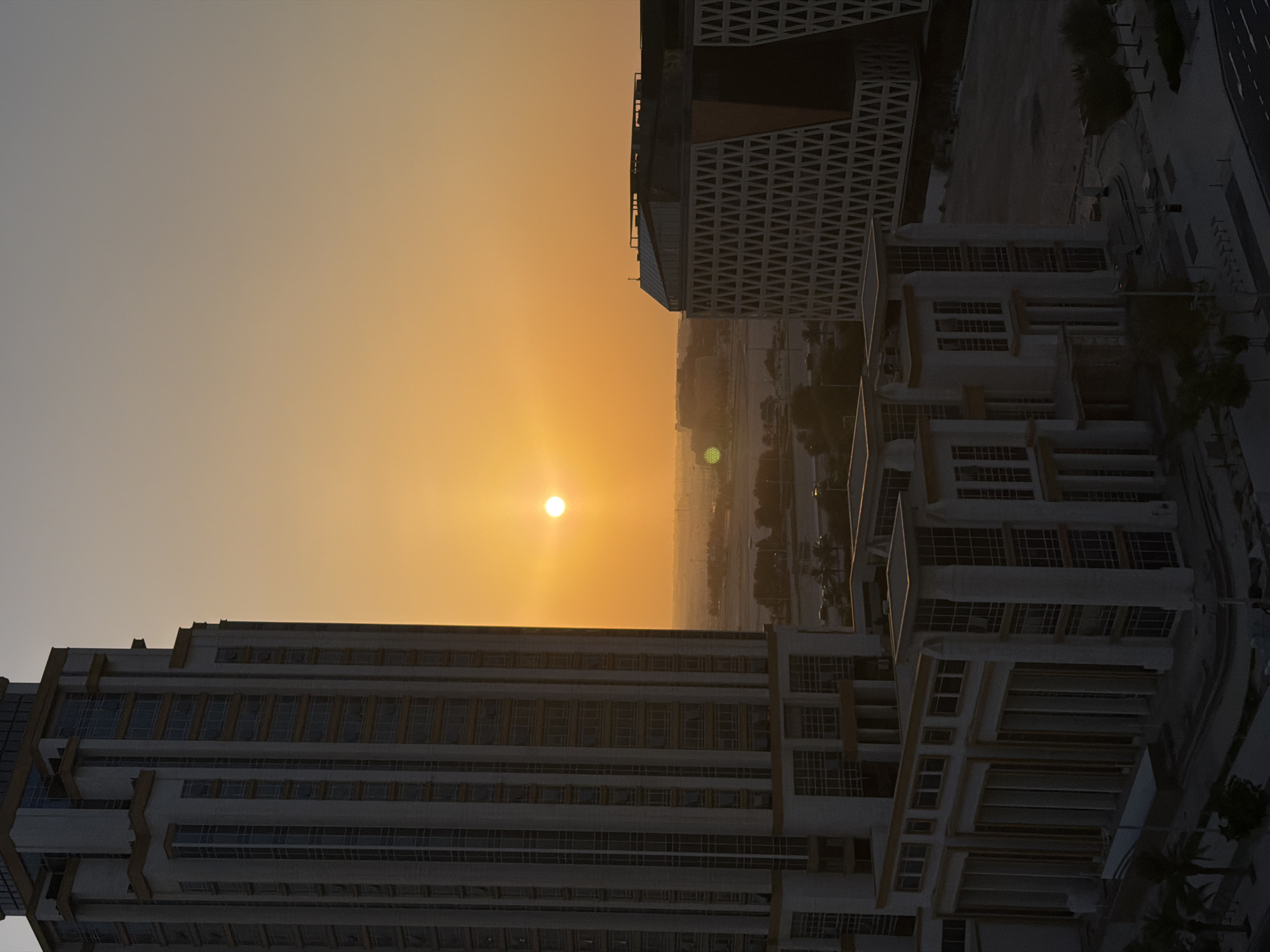 The image depicts an aerial view of a modern urban landscape at sunset. The sky is a warm gradient of orange and yellow, with the sun a bright, luminous orb near the center-left. The sun's rays illuminate the scene, casting a soft glow over the buildings and creating a hazy atmosphere in the distance.
Prominently featured are several large, multi-story buildings with contemporary architectural designs. One building in the foreground, on the left, is long and rectangular with a facade of numerous windows and horizontal banding. Another building, to the right and slightly further back, features a more intricate design with a patterned facade and multiple levels. In the background, more structures and possibly a city skyline are visible, shrouded in the sunset haze.
The image captures a moment of transition, likely the end of a day, with the natural light of the setting sun dominating the scene. The overall impression is one of a developed, perhaps business-oriented or residential, urban environment bathed in the golden light of dusk. There are no visible people, and the focus is on the architecture and the atmospheric quality of the sunset. The location is not specified, but the style of architecture suggests a modern city, possibly in a desert climate given the warm, hazy sky. The image evokes a sense of calm and grandeur associated with sunsets over developed areas.