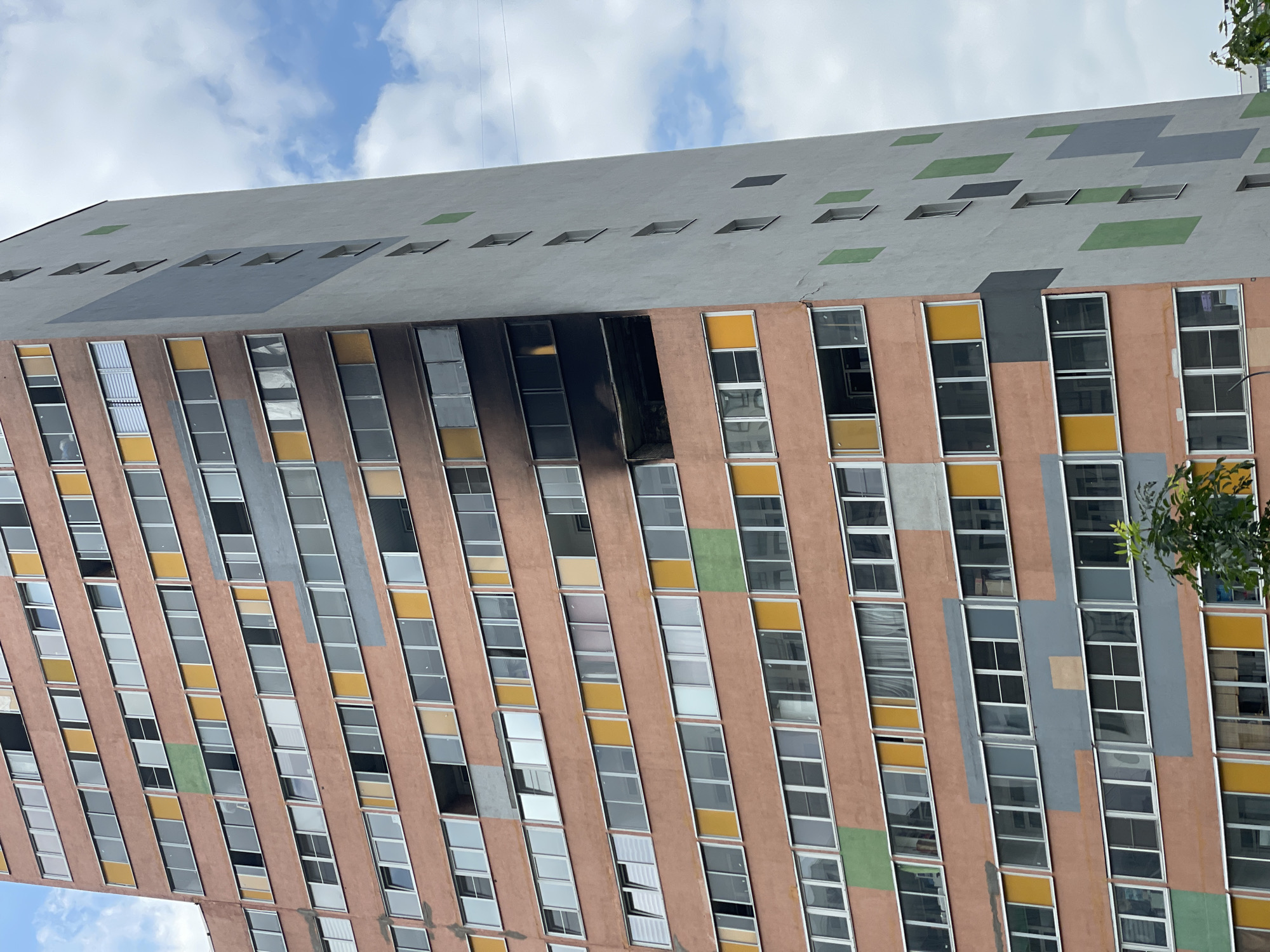 The image shows a tall, multi-story apartment building with a segment of the facade blackened by fire. The building has a reddish-orange main facade with various colored window panels, including yellow and green. The right side of the building is a lighter gray with large, abstract geometric shapes in gray and green. Several windows are visible across the building, some open and some closed. The fire damage is concentrated on several floors in the middle section of the building, leaving charred remnants of windows and balconies. The sky above is partly cloudy with white clouds against a blue background. The lower right corner shows a glimpse of green foliage from a tree. The context provided indicates this scene is in Murino, Russia.