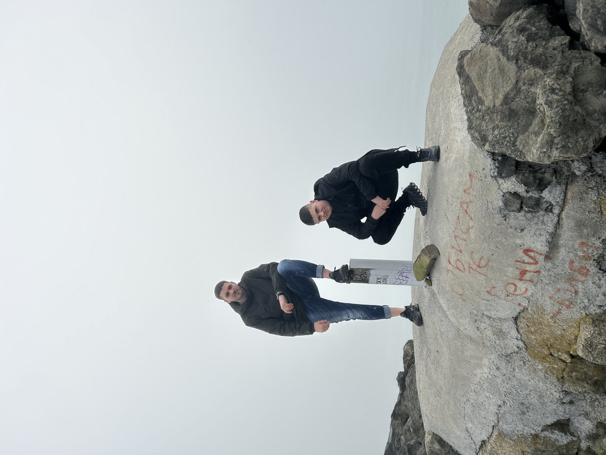 Two men are posing on a concrete structure next to a body of water. The man at the top of the frame is crouched down, wearing a black jacket and pants. The other man is standing with one foot on a pole, wearing a dark jacket and blue jeans. The location is the city of Unknown, Unknown.