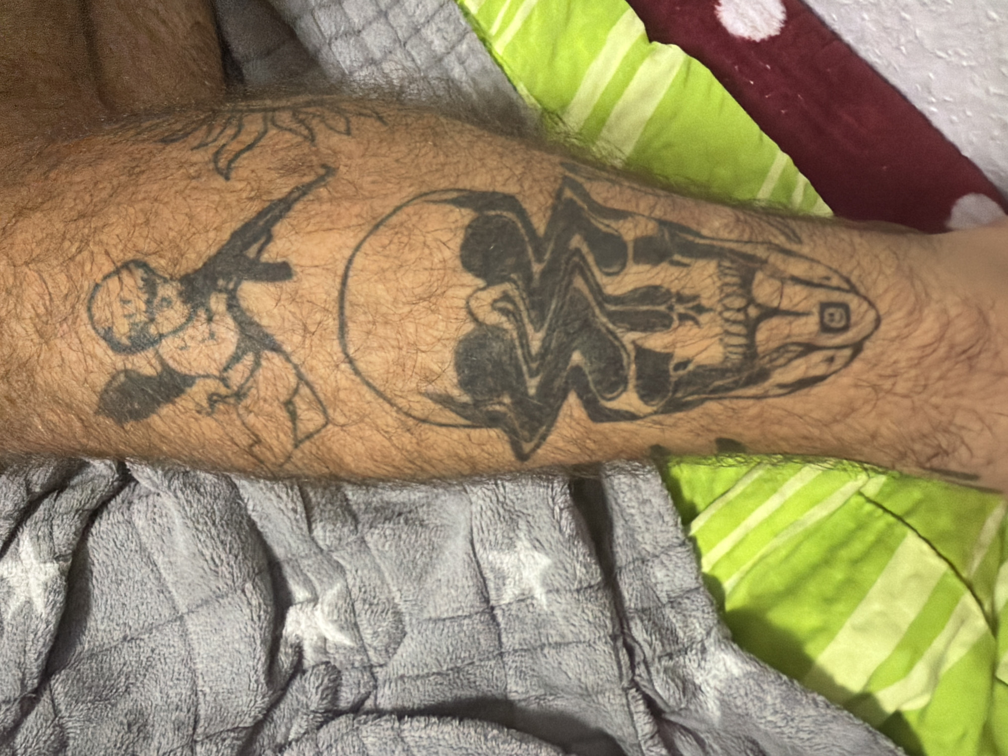 The image shows a close-up of a person's leg with tattoos. One tattoo is a skull with a distorted, wavy design on its face. Another tattoo above it appears to be a cherub holding a weapon. The leg is covered in dark hair. The person is lying on a gray, quilted blanket and a green and white striped fabric. A dark red object with white polka dots is also visible. The scene does not contain any elements that would allow for specific location identification within Dorsten, Germany.