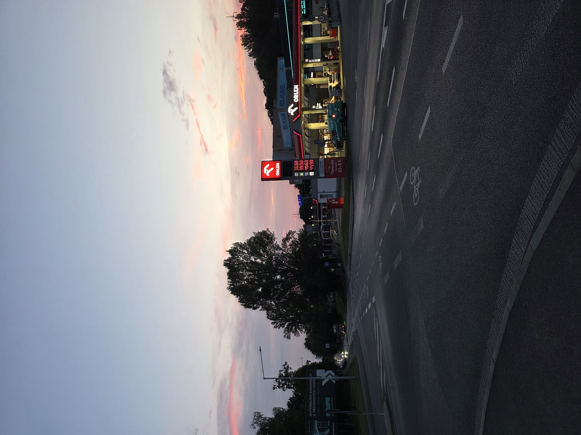 A gas station with the Orlen brand is visible on the right side of the image, with red and white signage illuminated against a dusk sky. Several cars are parked at the pumps, and a green van is driving through. The gas station is located at a street intersection with traffic lights and road markings, including a bicycle lane symbol. A large tree stands to the left of the gas station, and a billboard is visible further to the left. The sky is filled with streaks of pink and orange clouds, suggesting a sunset or sunrise. The overall scene depicts a quiet evening at a roadside gas station.