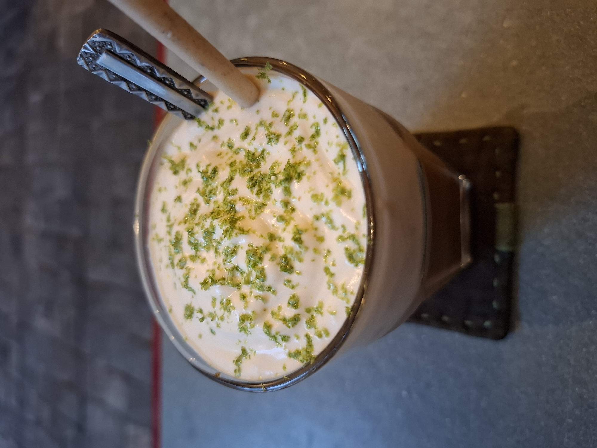 A close-up, overhead shot shows a beverage in a clear glass. The drink is topped with a generous layer of white foam, sprinkled with bright green zest. A light brown straw and a silver spoon with a decorative handle are inserted into the foam. The glass sits on a square, dark brown coaster with stitching around the edges. The background is blurred, appearing to be a textured, gray surface with hints of red and blue. The setting is likely a cafe or restaurant in Nha Trang Ward, Vietnam.