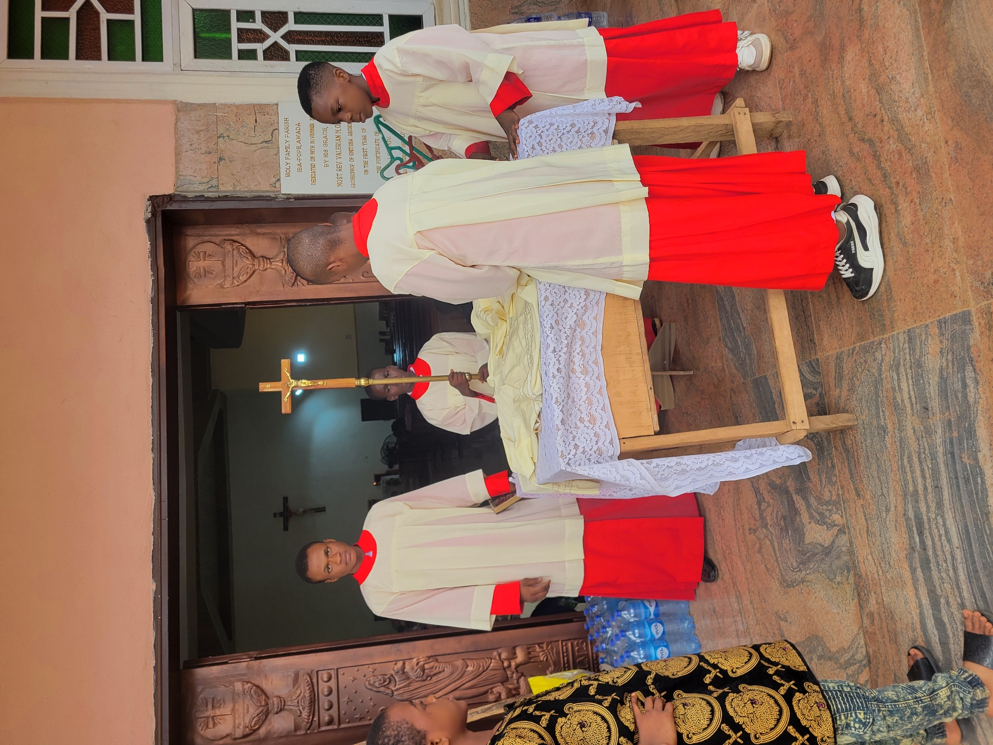 Here's a detailed description of the image:
**Scene:** The photo captures a scene outside the Holy Family Parish in the Oba-Pope area of Awada, Obosi, Nigeria. A plaque near the entrance indicates that the church was dedicated on November 16th.
**People:** Several altar servers are present, dressed in matching uniforms: cream-colored robes with red accents. One server stands in the foreground, looking toward the camera, while another is holding a processional cross inside the doorway. Two other servers are near a wooden table draped with a white lace cloth. One person is partially visible in the bottom foreground wearing a patterned garment.
**Objects:** Key objects in the image include the wooden table covered with a white lace cloth, a processional cross, and several bottles of water on the ground. The entrance door is ornately carved with religious symbols.
**Actions/Interactions:** Some servers are arranging or attending to the cloth on the table. Another appears to be carrying the processional cross into or out of the church.
**Environment:** The location is outdoors, with what appears to be tiled or stone flooring. A window with decorative panes can be seen above the doorway.