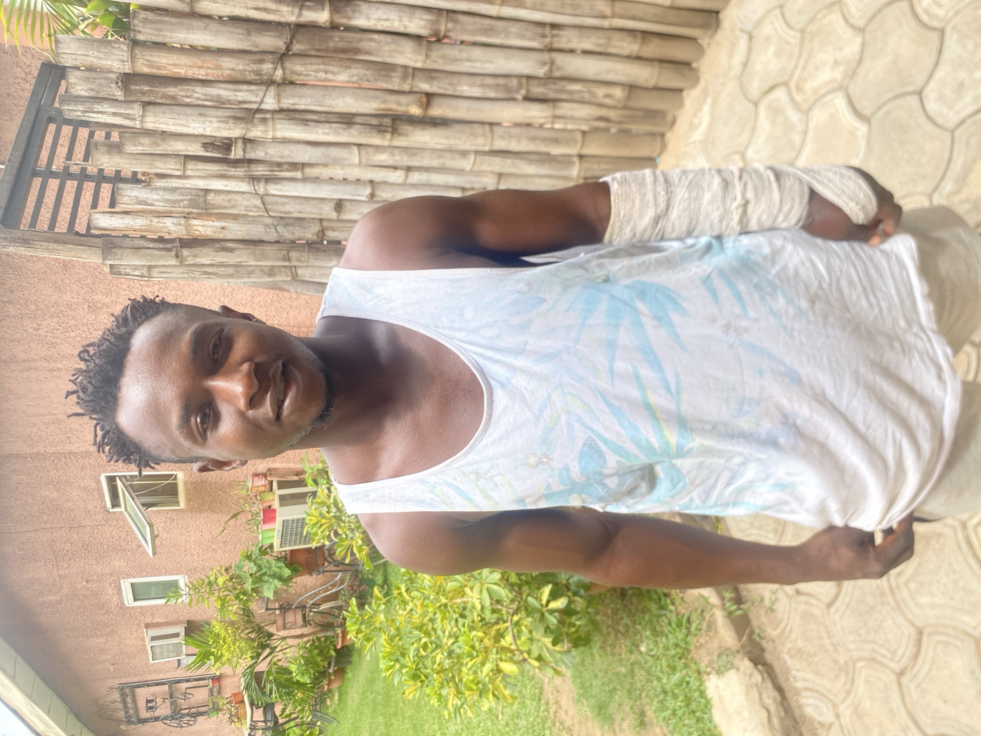 The image features a man with dark skin and short dreadlocks, wearing a white tank top with a light blue leaf pattern and beige shorts. His left arm is bandaged from the mid-forearm to his hand. He is standing outside, partially in front of a wall constructed of horizontal bamboo-like material. Behind him to the left is a building with multiple windows, one of which is open. In front of the building is foliage. The ground is paved with interlocking tiles. The scene appears to be set in daylight. The exact location in Barden Yana, Nigeria, cannot be determined from the information in the image.