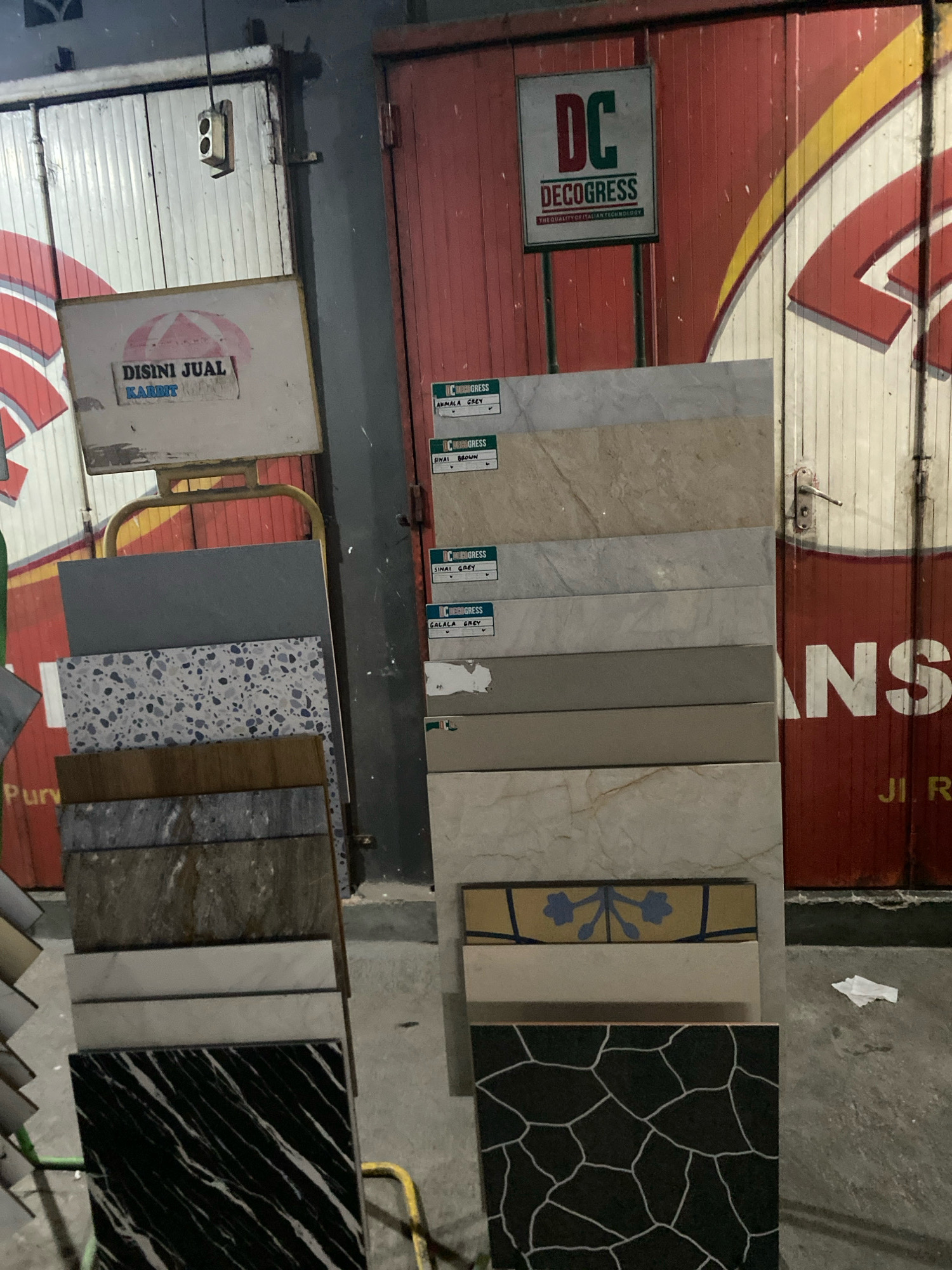 This image depicts an outdoor retail display of tiles. In the foreground, a rack showcases a variety of ceramic tiles with different patterns and colors, including marble-like, speckled, and geometric designs. To the right, a stack of larger tiles are arranged vertically. A sign reading "DISINI JUAL KARET" (meaning "Rubber Sold Here") is visible on the left, with a logo of a red and white umbrella. Above the tiles, a sign with "DC DECOGRESS" and the tagline "THE QUALITY OF ITALIAN TECHNOLOGY" is mounted on a red corrugated metal door. The background features closed rolling doors and a sign that appears to read "JI. R". The scene is likely in Kertek, Indonesia, given the context provided. The lighting suggests it might be evening or nighttime.