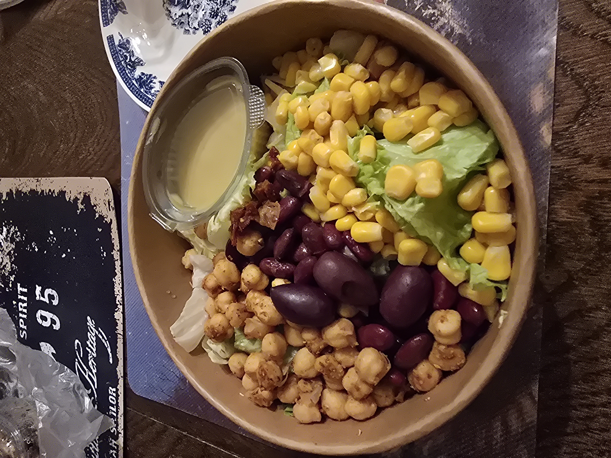 The image shows a bowl of salad on a table. The salad contains lettuce, corn, various beans (including kidney beans and possibly black beans), fried chickpeas, and sun-dried tomatoes. A small container of dressing is placed in the bowl. The bowl appears to be made of cardboard or a similar material. The table is made of wood. A blue and white plate is visible in the upper left corner of the image, and a black and white sign with the words "Spirit 95 Heritage Sailor" is visible to the left of the bowl. The presence of a sign with the word "Sailor" suggests a nautical theme. While Pernik is a city in Bulgaria, the image provides no specific details to confirm if the location is Pernik.