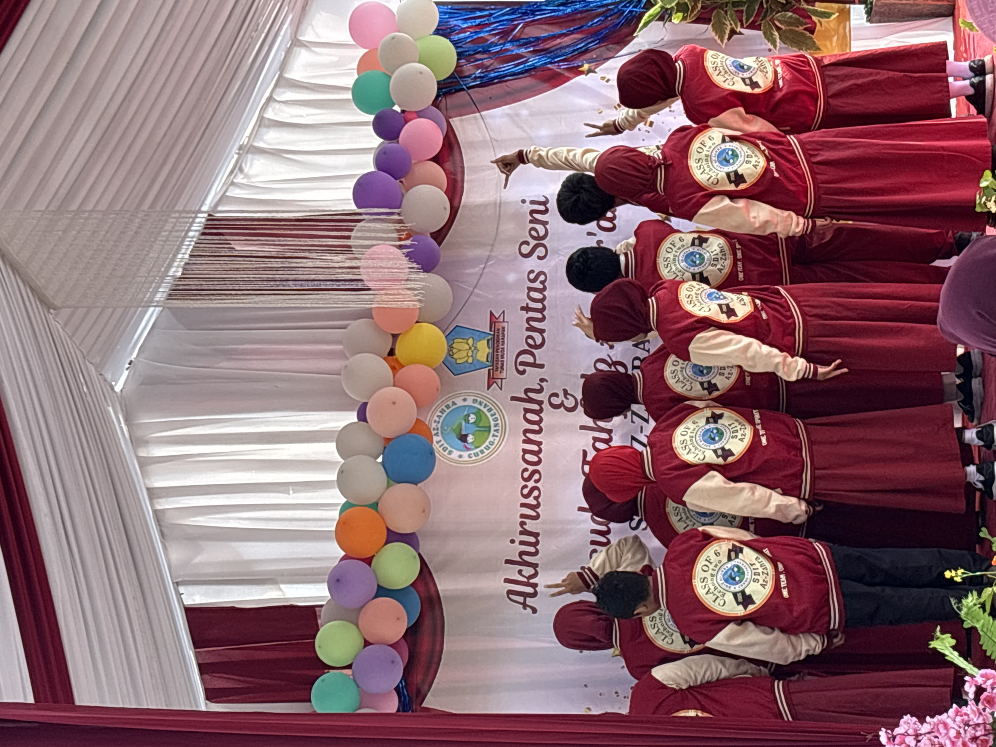 In Sukabakti, Indonesia, a group of students, primarily wearing maroon hijab and matching skirts, stand facing away from the camera. They are adorned with red varsity jackets featuring cream-colored sleeves and embroidered circular logos on the back, which read "CLASS OF 6" and "SDIT AZ-ZAHRA". Some students are raising their hands in the air, with one making a peace sign and another forming a lasso gesture. A banner behind them reads "Akhirussanah, Pentas Seni & Yudisium Fakh... 2027-2023", indicating a graduation and arts performance event. The backdrop is a draped white and maroon fabric, and a cluster of colorful balloons, including green, yellow, purple, orange, and pink, floats above. Decorative white string fringe hangs from the ceiling structure. The setting appears to be an indoor or covered outdoor stage.
