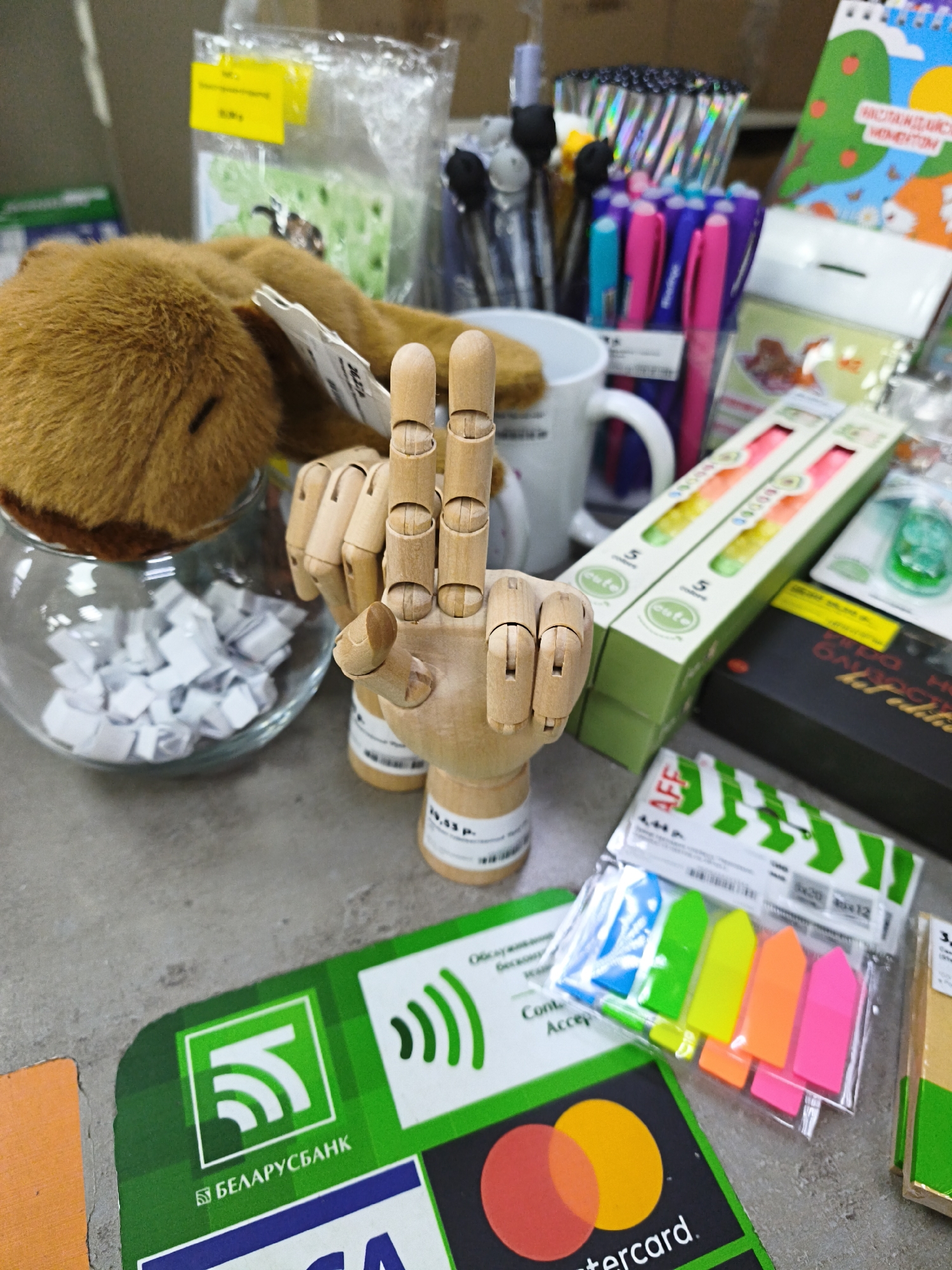 The image depicts a collection of stationery and office supplies arranged on a grey surface, possibly a table or counter. In the foreground, a wooden mannequin hand is posed with two fingers extended in a peace sign. To the left of the hand, a brown stuffed toy, resembling a rabbit, lies partially obscured. A glass bowl filled with small white folded papers is situated behind the toy.
Dominating the middle ground are several items: a white mug, a collection of colorful pens (pink, blue, and purple), and two green boxes containing what appear to be candles or long cylindrical items, each with the text "safe" visible. Further to the right, a portion of a colorful children's notebook with an illustration of a hill and animals is visible.
In the background, several packages and labels are present. A yellow price tag is affixed to a clear plastic bag containing green paper. Below the mannequin hand, a large green sticker with white icons and the text "БЕЛАРУСБАНК" (Belarusbank) is prominent. Next to it, another sticker displays the Mastercard logo. A package of brightly colored sticky notes (pink, orange, yellow, green, and blue) is also visible.
The scene suggests a retail or office environment. The presence of the "БЕЛАРУСБАНК" sticker strongly indicates the location is in Belarus, likely a point-of-sale or promotional display within or near a bank branch, or a store that accepts Belarusbank services. Given the specific mention of Belarusbank, a location within the city of Minsk is plausible. The overall impression is one of an organized display of various small items. No people are present in the image.