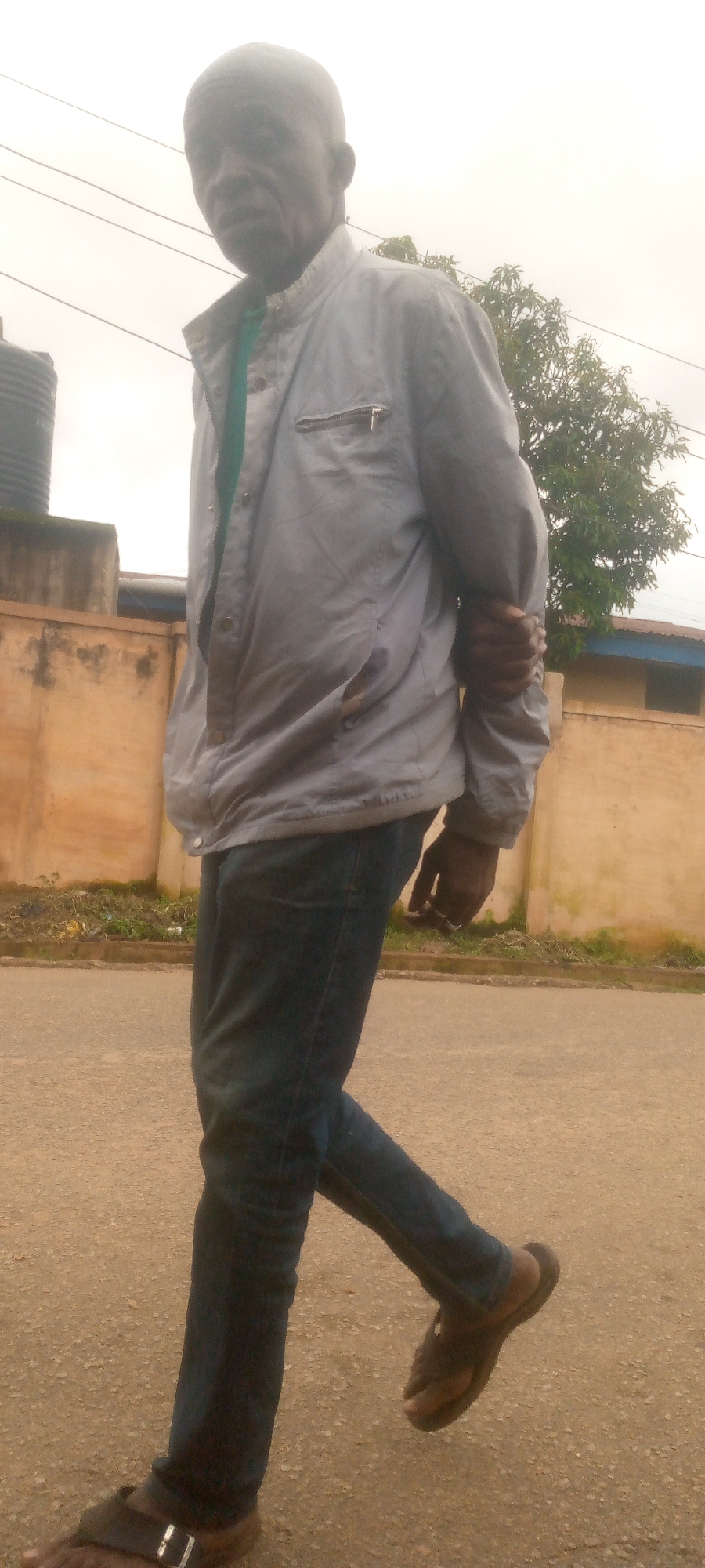A full-body, medium shot captures a Black man walking on a paved surface in what appears to be a residential area. The man is wearing a light blue jacket with a zipper pocket on the left chest, a green shirt underneath, dark blue jeans, and dark brown sandals. His left hand is in his pocket, and his right arm is bent with his hand gripping his left arm. The background features a beige building with a blue-capped roof, a large black water tank, and a tree. The sky is overcast, suggesting cloudy weather. The man is positioned in the center of the frame, walking towards the left.