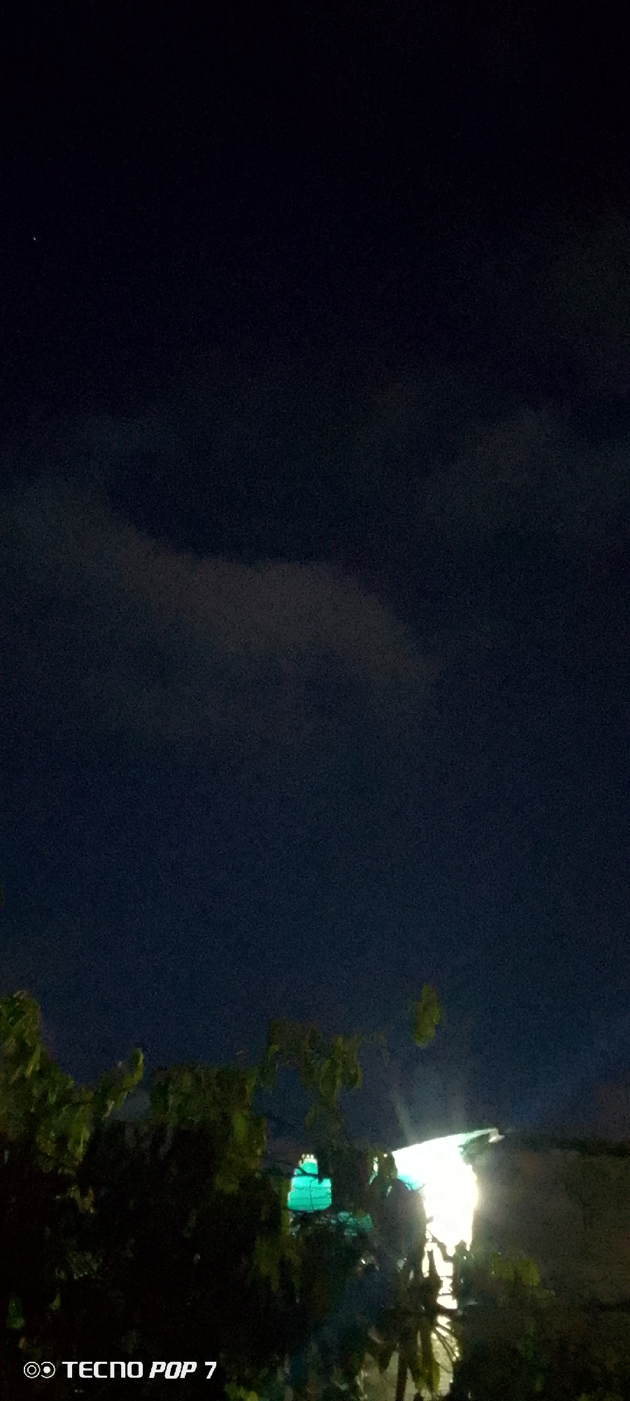 This is a nighttime photograph taken in Altagracia, Venezuela. The image depicts a dark sky with some faint clouds. In the foreground, there are lush green trees and foliage, partially obscuring the view of the background. To the right of the center, a bright light source illuminates the scene, possibly from a building or streetlamp, casting a strong glow upwards and to the right. To the left of the bright light, a small structure with green lighting is visible, appearing to be a decorative element or a small building. The bottom left corner of the image contains a watermark that reads "TECNO POP 7". No people are visible in the frame.