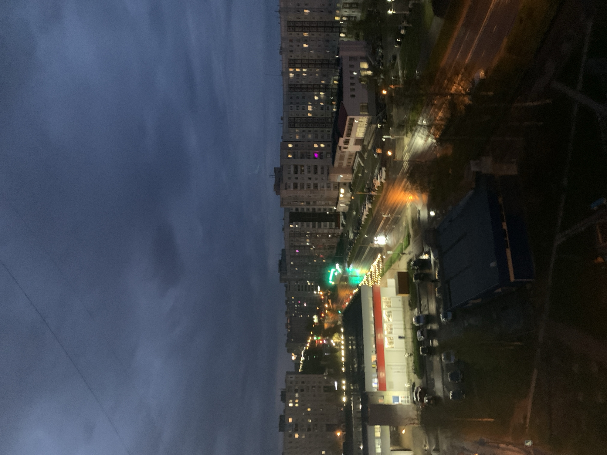 The image shows a view from a high vantage point, possibly a window, overlooking a city at dusk or night. The location is Severodvinsk, Russia, inferred from details such as the type of architecture.
The left side of the image shows a dark, cloudy sky. The right side depicts a cityscape. Rows of apartment buildings with illuminated windows line the street. A street runs between the buildings, illuminated by streetlights and the headlights of moving vehicles. Further away, a building appears to be a shopping center. A parking lot filled with cars is visible in front of the building.