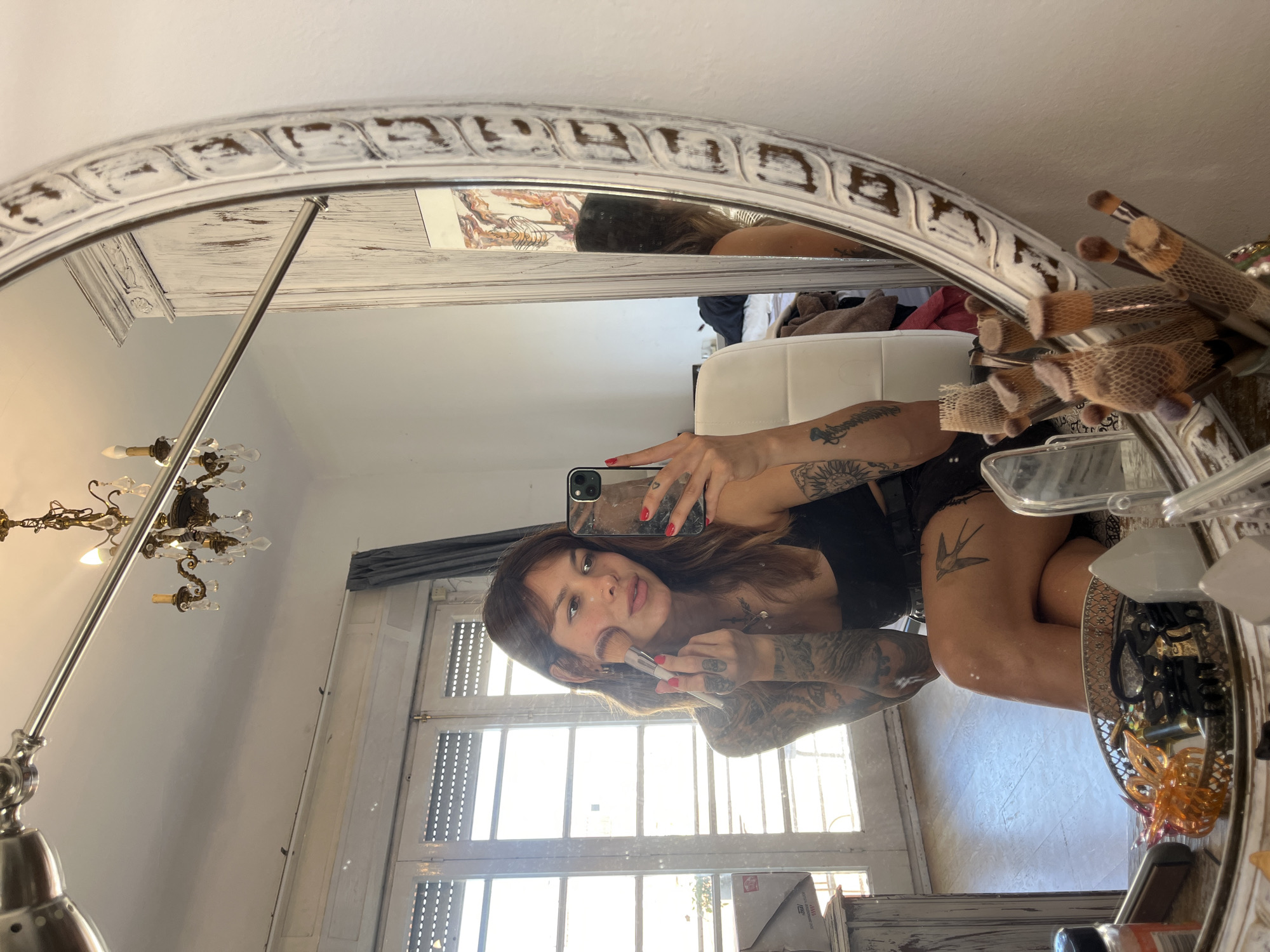 A person is applying makeup in front of a mirror in Barcelona, Spain. The person is wearing a black top and shorts and has tattoos on their arms and legs. They are holding a makeup brush to their cheek and looking at their reflection in the mirror. The mirror is ornate with a white distressed frame. In the reflection, a chandelier is visible hanging from the ceiling, and a large window with blinds is to the left. A chair and a dresser with various items on it, including makeup brushes, are also reflected in the mirror. The lighting suggests it is daytime, with sunlight coming through the window.