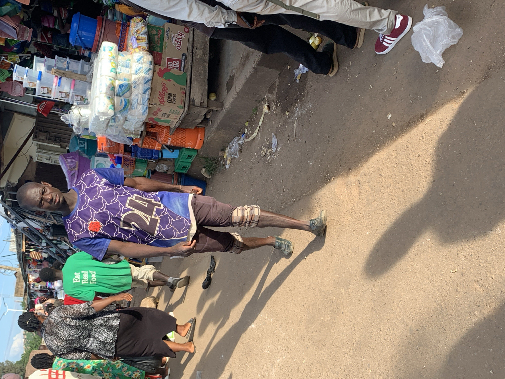 An outdoor market scene unfolds in Jos, Nigeria, with several individuals navigating a dusty pathway lined with stalls. In the foreground, a man in a purple shirt with a white graphic and the number "24" walks towards the viewer. He wears dark brown, knee-length shorts with checkered patterns around the knees and dark, lace-up shoes. To his left, a woman in a black skirt and a patterned grey and white blouse walks away from the viewer, carrying a dark bag. Behind her, a man in a bright green t-shirt with the text "Eat Real Food" printed on the back walks away, wearing light-colored shorts and dark sandals.
The background is dominated by market stalls filled with various goods. Stacks of plastic containers, paper towel rolls, and boxes of cereal are visible. Orange propane tanks are also present among the merchandise. The ground is a mixture of packed dirt and some scattered debris, including plastic bags and what appears to be discarded food waste. The overall atmosphere suggests a bustling, active marketplace under natural daylight.