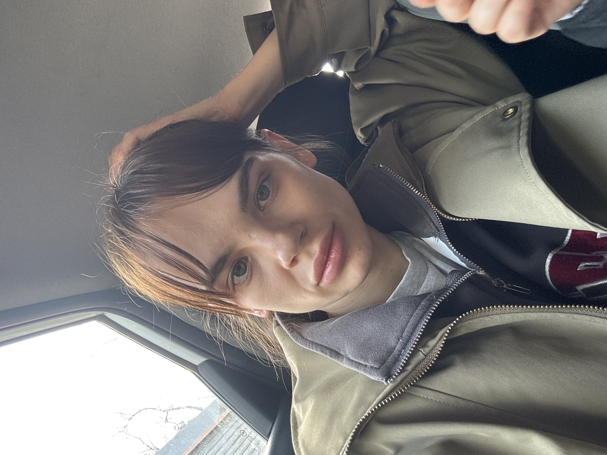 A person is inside a vehicle, possibly a car, as suggested by the window in the lower left corner showing a blurred view of a building and tree branches. The person is wearing a khaki-colored jacket with a zipper, layered over what appears to be a gray hooded sweatshirt and a T-shirt. The person has light skin, dark brown hair with bangs, and appears to be looking directly at the camera. Their left hand is touching their hair. There is no contextual information in the image to determine if the location is in Rostov-on-Don, Russia.