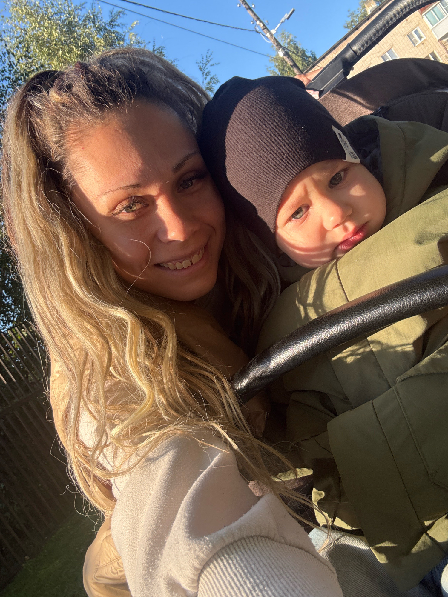 In this close-up, outdoor photograph taken in the city of Rusanovo, Russia, a woman with long, wavy blonde hair smiles at the camera. She is wearing a light-colored sweatshirt and is positioned to the left of the frame. To her right, a baby wearing a dark beanie and a green jacket is visible, looking towards the camera with a neutral expression. The baby is nestled in a stroller, the handle of which is prominently featured in the foreground. The background includes a bright blue sky, some trees, a fence, and a building with windows. The lighting suggests it is daytime, possibly late afternoon, with sunlight casting shadows on the subjects.