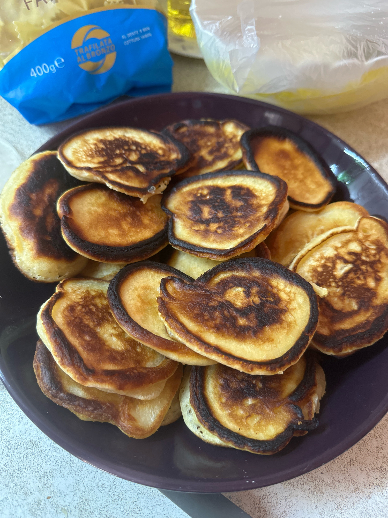 A dark purple plate is filled with a stack of small, round pancakes. Many of the pancakes are slightly overcooked, with dark brown or black edges. Some appear to have unevenly cooked centers, with lighter golden-brown patches. In the background, out of focus, are two bags. The bag on the left is blue and has "TRAFILATA AL BRONZO" and "400ge" printed on it. The bag on the right is clear plastic and contains a yellow substance. The surface the plate rests on appears to be a light-colored speckled countertop. The scene is indoors, likely a kitchen. There is no indication of the city of Kalininskaya, Russia.