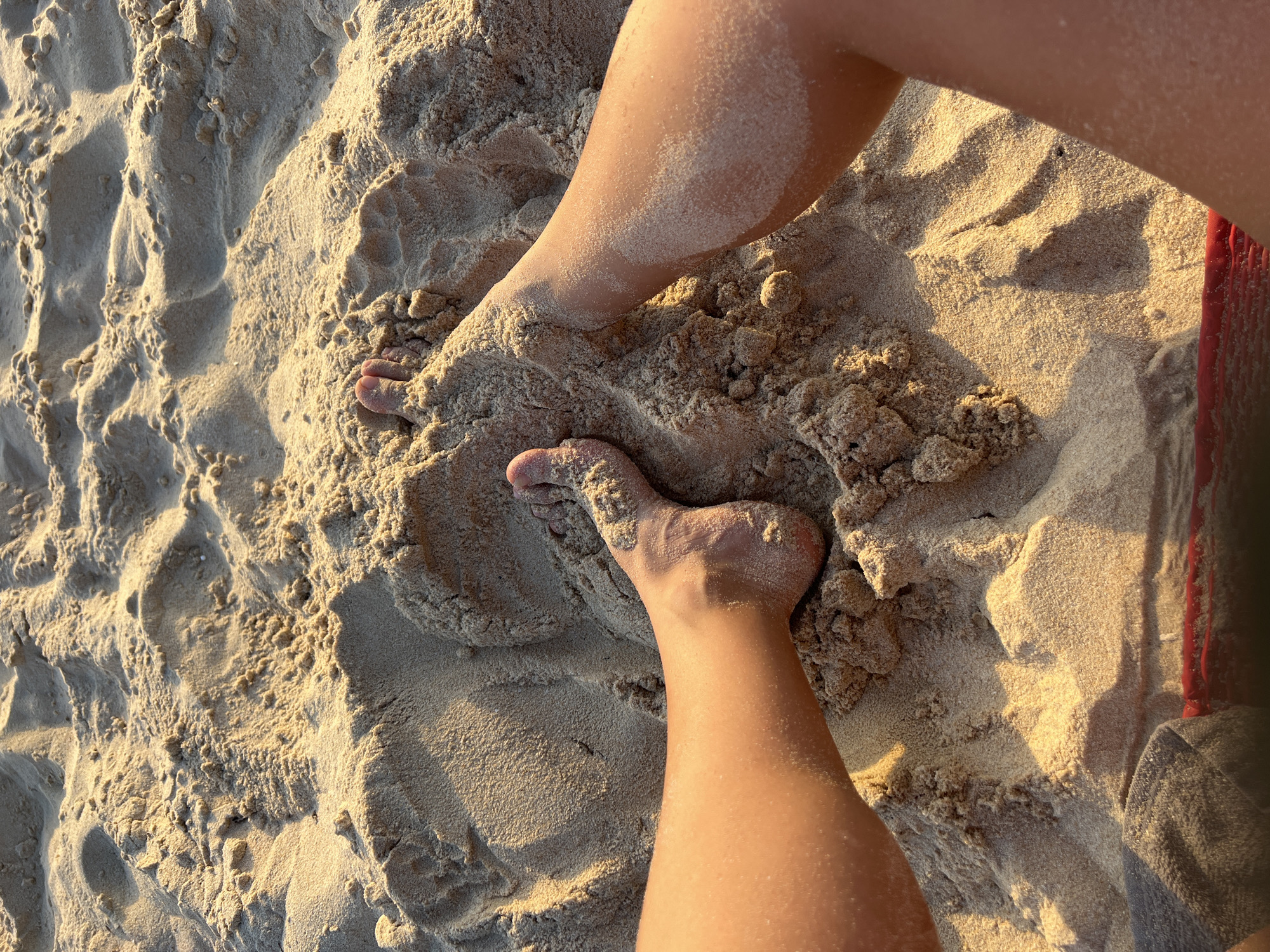 A close-up, overhead view of two bare feet partially buried in damp, golden sand. The sand is textured with ripples and small clumps, suggesting it has been recently disturbed. The skin of the feet and legs is tanned and dusted with fine grains of sand. Sunlight casts long shadows across the sand, highlighting the uneven surface. The scene is set on a beach in Choeng Thale, Thailand.