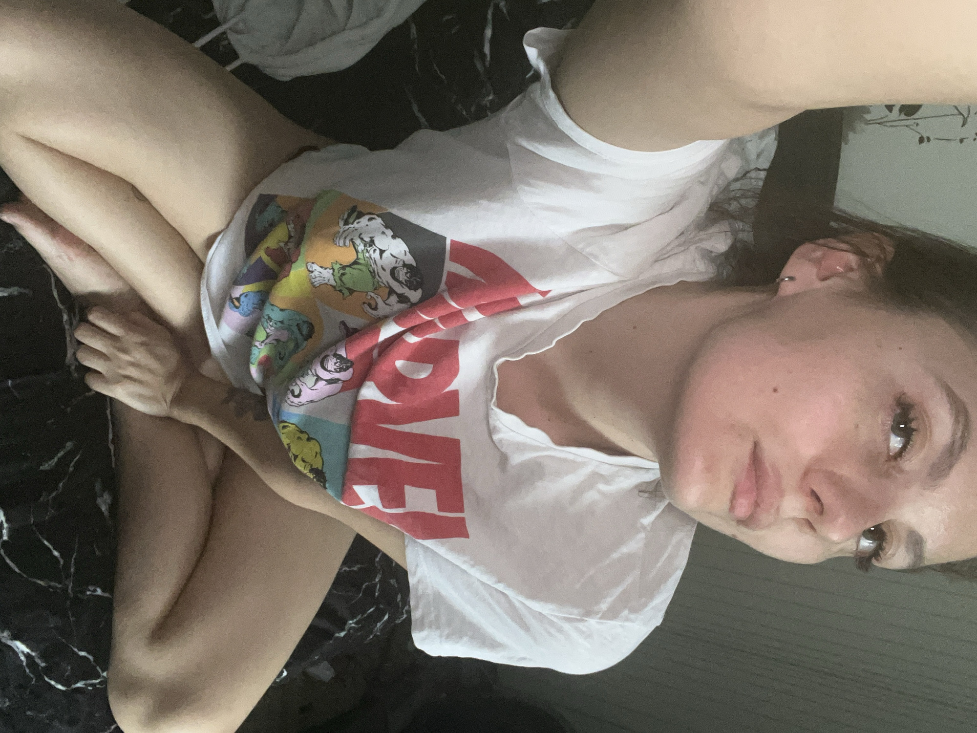 A young woman is captured in a selfie from an overhead, slightly tilted perspective, as she lies in a relaxed position indoors. She has dark hair and fair skin, with her face, including her eyes, nose, and mouth, visible as she looks towards the camera with a neutral expression. She is wearing a white short-sleeved t-shirt featuring a colorful graphic design of comic book characters, notably including what appears to be the Hulk. Prominently displayed on the shirt is the word "LOVE" in large red capital letters. Her legs are bent, and her feet are crossed, suggesting she is lounging on a surface. This surface is dark, with white, marble-like patterns, possibly indicating a bedspread or bedding. The background is soft and indistinct, showing a light-colored wall or surface. The lighting is diffused, consistent with an indoor setting, and no specific time of day is discernible. She wears earrings in one ear.