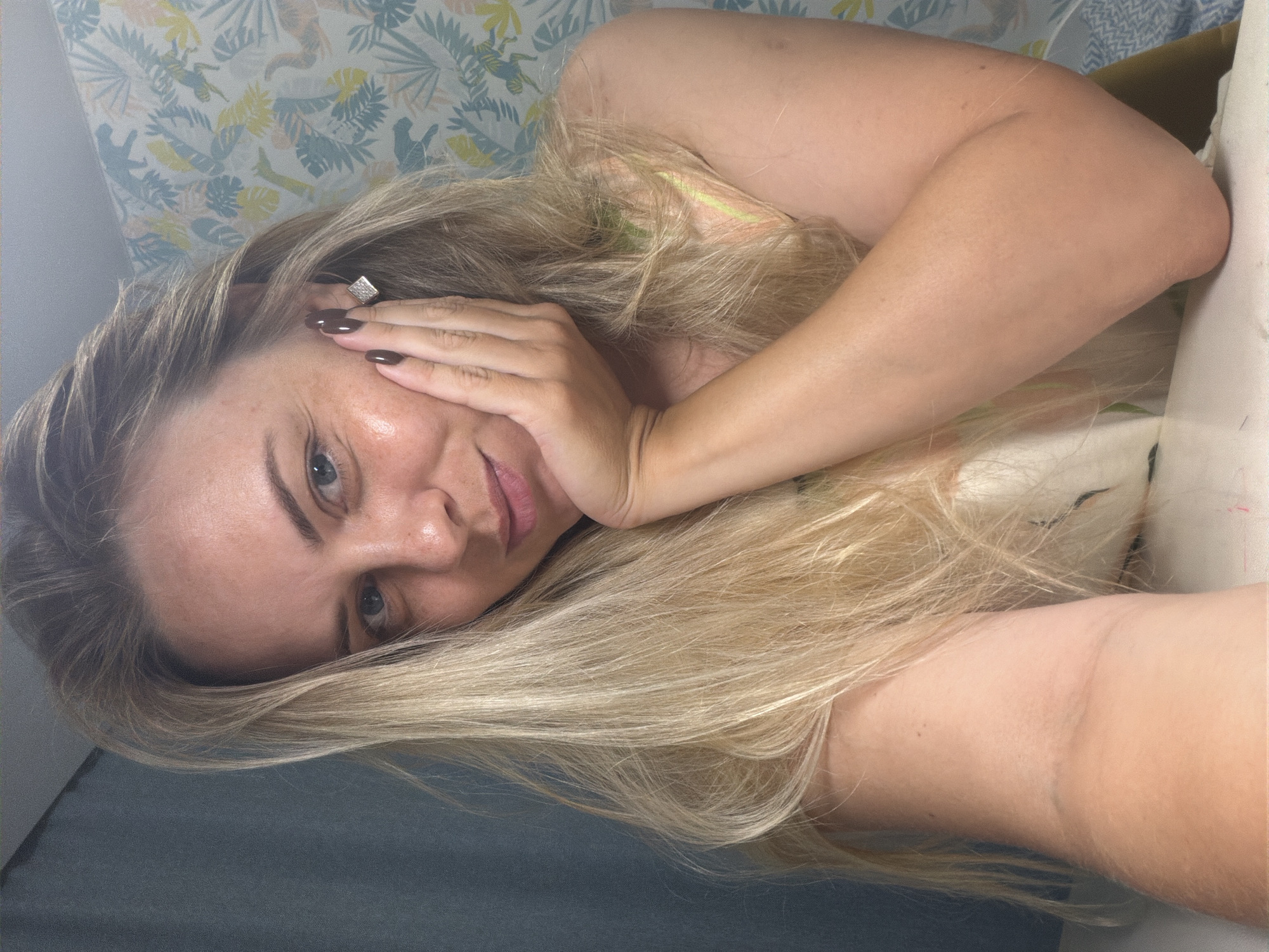 This is a selfie of a woman with long, blonde hair, lying on her side. Her left hand is resting on her forehead, obscuring part of her face. She is wearing a swimsuit with thin straps. The background appears to be a bedroom with a patterned wallpaper and a grey surface that could be a duvet or another piece of bedding. The lighting suggests it is daytime, possibly indoors. The woman's expression is relaxed, and the overall mood is casual and intimate. No text is visible in the image. The scene is likely set in Votkinsk, Russia, based on the provided location context.