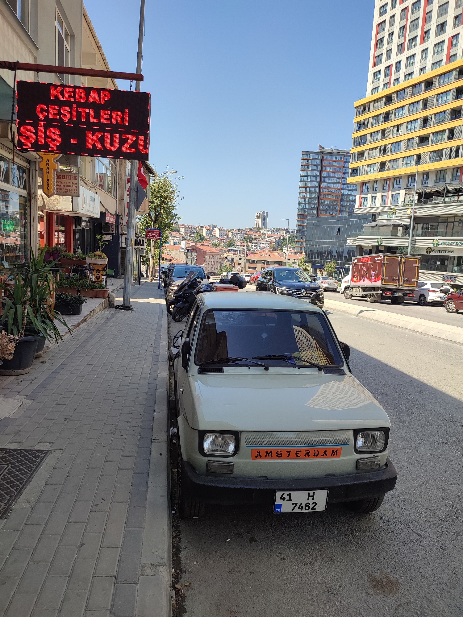 The image captures a street scene in Kadıköy, Turkey, on a bright, sunny day. In the foreground, a light-colored, older model car is parked along the side of the road, facing away from the viewer. It has a black bumper with a license plate that reads "41 H 7462 TR". An orange sticker with the word "AMSTERDAM" is visible on its front.
To the left of the car, a sidewalk is paved with grey rectangular tiles. A red LED sign hangs above a storefront, displaying Turkish text that appears to advertise food items like "KEBAB ÇEŞİTLERİ" (kebab varieties) and "ŞİŞ - KUZU" (skewer - lamb). Various plants in black pots are placed along the sidewalk in front of the shops.
Further down the street, several other vehicles are parked or in motion, including a dark SUV, a scooter, and a white truck with a brown cargo box. In the background, modern high-rise buildings with yellow and red accents rise above older, lower-rise residential buildings with red-tiled roofs, typical of a Turkish urban landscape. The sky is clear and blue. The overall impression is of a bustling city street with a mix of old and new architecture.