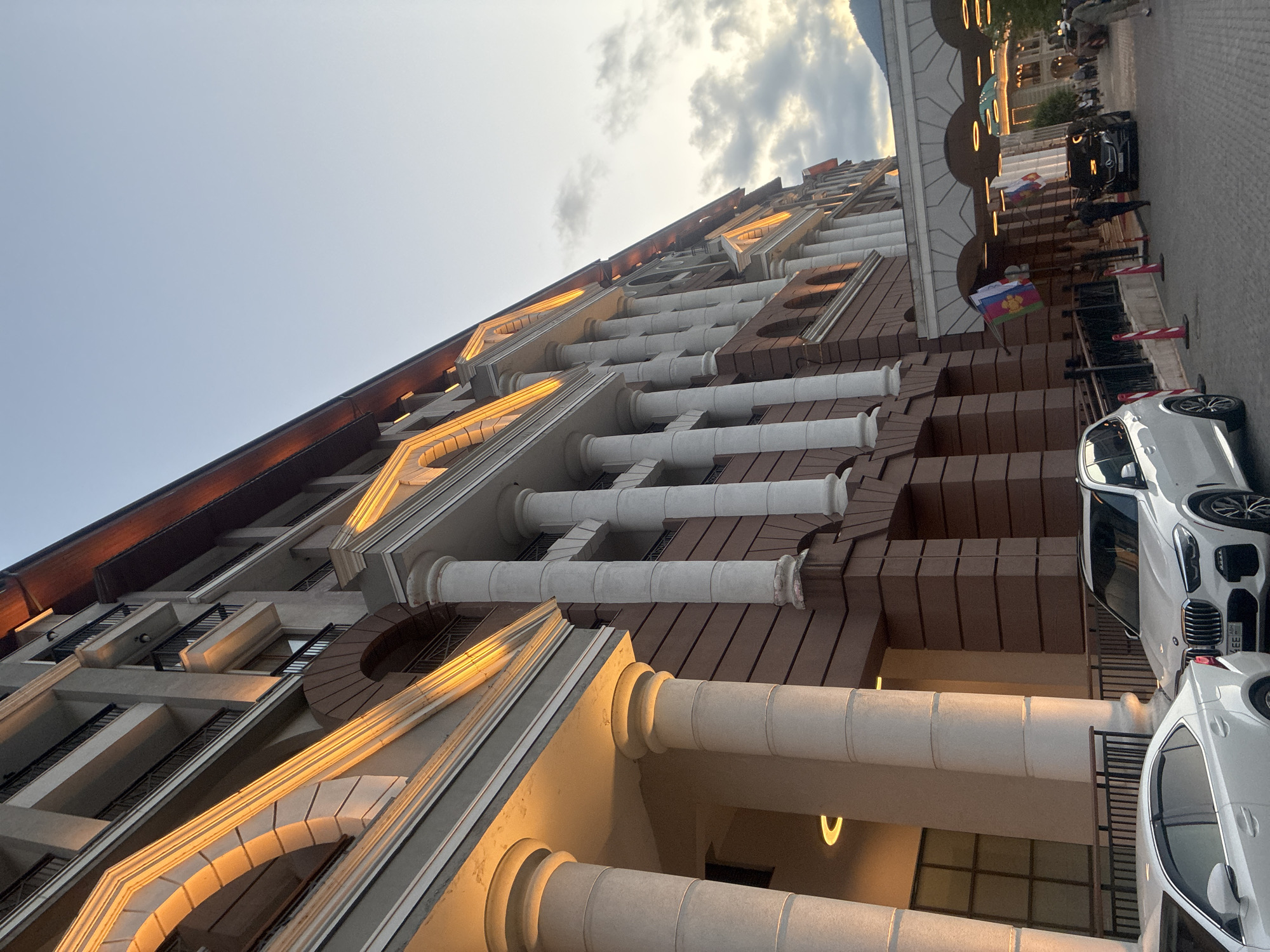 This image depicts the entrance to a grand hotel in Estosadok, Russia, characterized by its neoclassical architecture. The hotel features a facade with large, fluted columns and arched windows, illuminated by warm exterior lights that highlight the building's architectural details. A covered walkway with arches leads to the entrance, where two flags are prominently displayed.
In the foreground, two white cars are parked on a cobblestone street. The car on the right is a modern SUV, likely a BMW X5, identifiable by its grille and headlights. The car on the left appears to be a sedan. Several people are visible near the hotel entrance, with some individuals walking and others standing. One person is dressed in dark clothing and appears to be walking towards the hotel entrance. Another person in the background is wearing a light-colored uniform. The scene is set against a twilight sky with scattered clouds, suggesting it is either early morning or late evening. The overall impression is one of a well-maintained, upscale establishment in a scenic, possibly mountainous, region.
