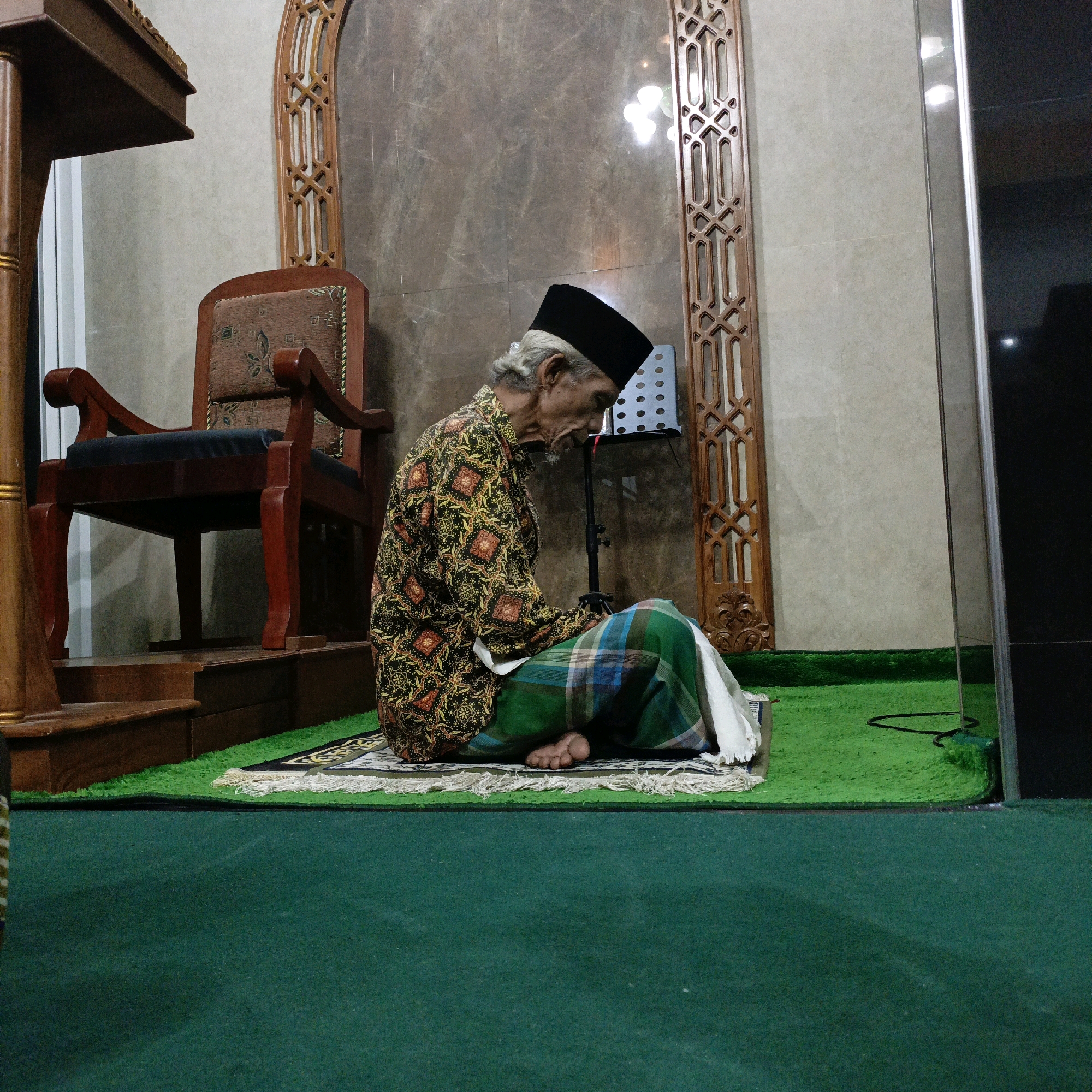 An elderly Indonesian man wearing a black peci hat and a batik shirt with a gold, black, and red pattern is seated in prayer on a rug. He is also wearing a green and blue checkered sarong and a white cloth. He is facing to his right, with his head bowed. He is sitting on a green carpeted area in what appears to be a mosque or prayer room. Behind him is a wooden chair with an ornate, upholstered seat, and to his left, a wooden structure with intricate carvings. The walls are tiled in a marble pattern. A music stand is visible behind the man. The foreground is dominated by a dark green carpet. The scene is lit by artificial lighting, creating reflections on the marble walls. This image was taken in Salatiga, Indonesia.