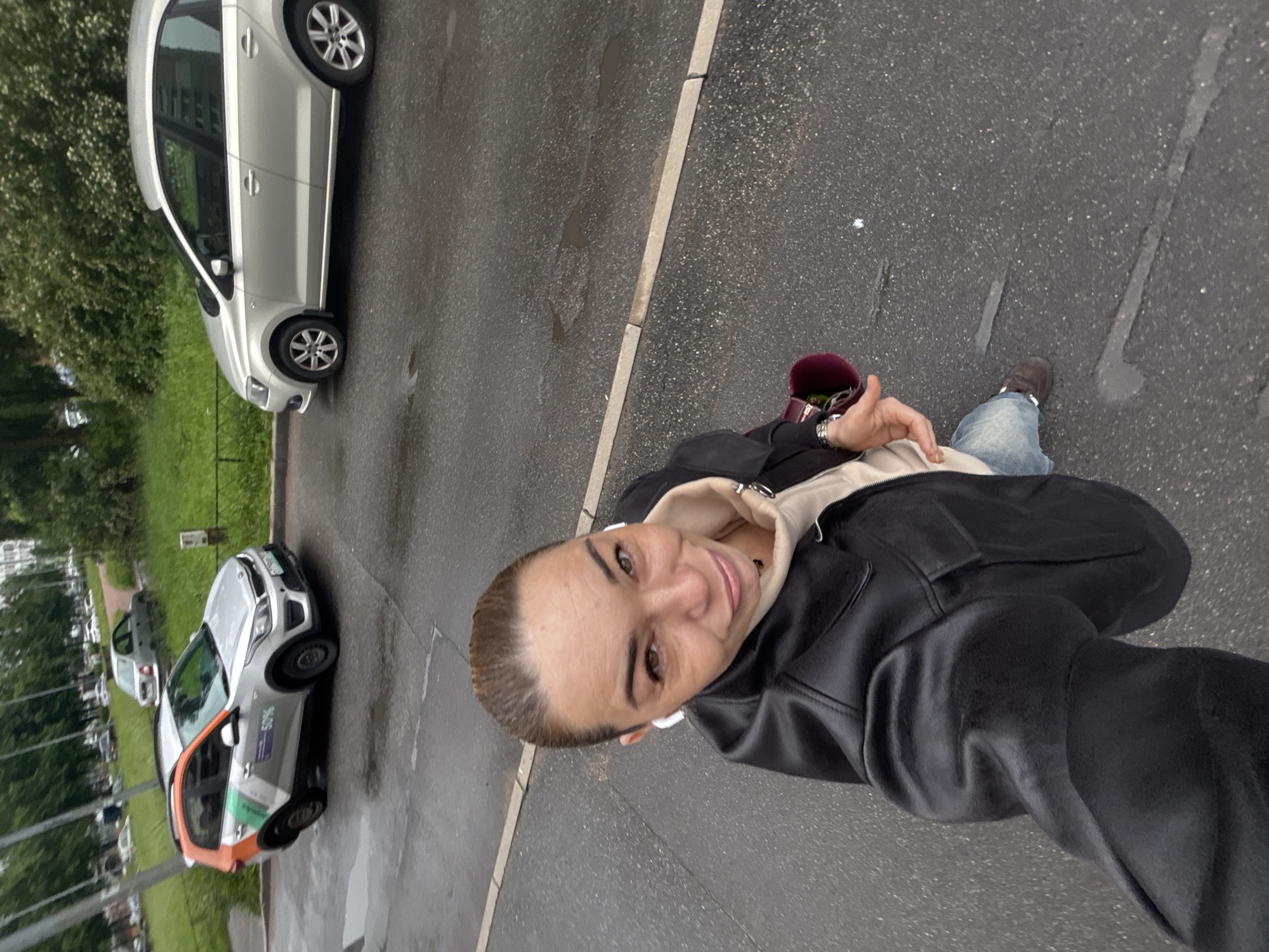 A woman with light brown hair pulled back stands on a wet asphalt street, looking directly at the camera. She is wearing a black leather jacket over a beige hoodie and blue jeans. White wireless earbuds are in her ears, and she holds a dark red bag in her left hand. In the background, on the right side of the image, a silver sedan is parked on the street. On the left side of the image, a silver and orange compact car with "50%" branding is parked. The street appears to be in a residential area, with trees and green grass bordering the road. The scene suggests an overcast day, with puddles of water visible on the asphalt. The location is Vsevolozhsk, Russia.