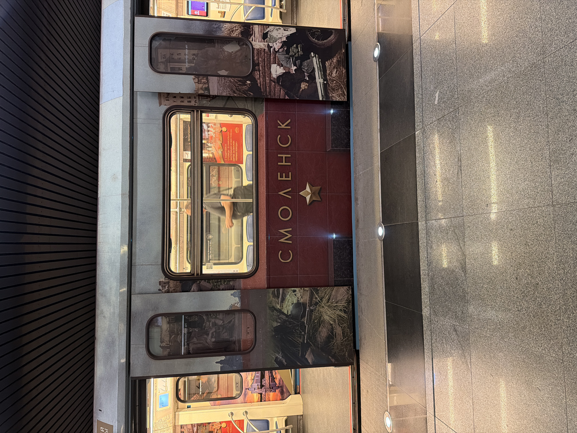 The image shows a side view of a Moscow Metro train carriage decorated with images and the word "СМОЛЕНСК" in Cyrillic. The carriage features historical wartime imagery. The interior of the carriage is visible through the windows. The train is at a station, with a tiled platform and wall visible to the right of the train. The picture is taken in Moscow, Russia.