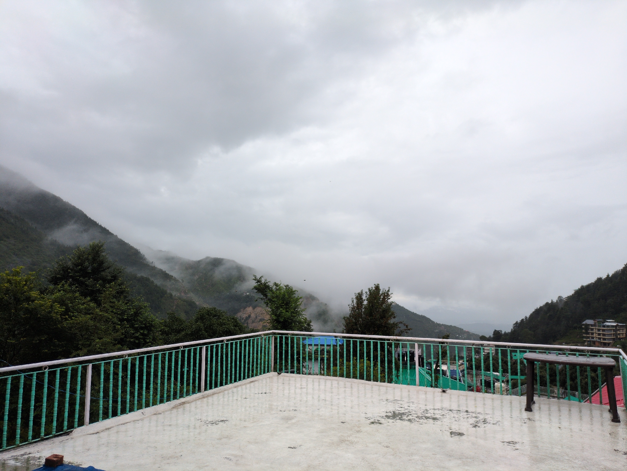 An overcast, possibly rainy day view from a balcony in Haini, India. The foreground features a white concrete balcony floor with puddles reflecting the sky and railing. A green metal railing with white accents borders the balcony. Beyond the railing, lush green trees cover sloping hills. In the distance, mist partially obscures the mountainous terrain and some buildings are visible, including a multi-story structure on the right side of the frame. A dark brown table with wooden legs stands on the balcony. The sky is filled with heavy, grey clouds.