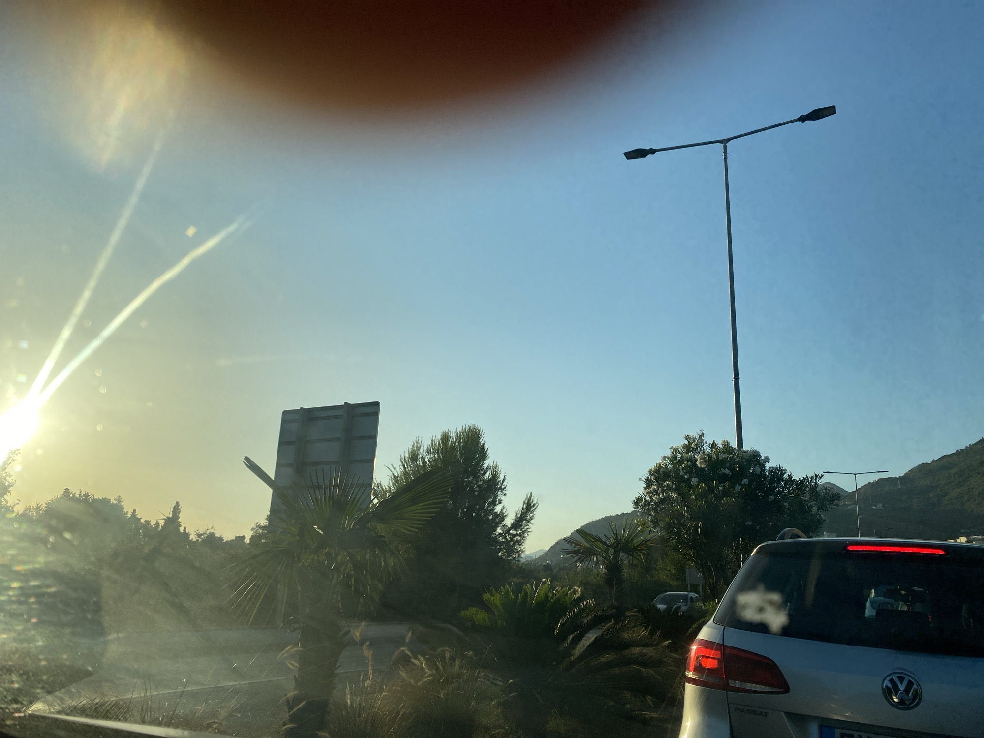 The image captures a view from within a vehicle, looking out through the windshield. The scene appears to be a roadside environment in Bar, Montenegro, indicated by the presence of lush Mediterranean vegetation, including palm trees and dense greenery. In the foreground, the rear of a silver Volkswagen Passat is visible, with its brake lights illuminated. Beyond the car, a traffic light pole with two lamps stands against a clear blue sky. Sunlight streams in from the left, creating lens flare and streaks across the image, suggesting it is either early morning or late afternoon. Further in the background, rolling hills are visible, covered in trees. The overall impression is one of a coastal town in a sunny climate, likely during daylight hours.