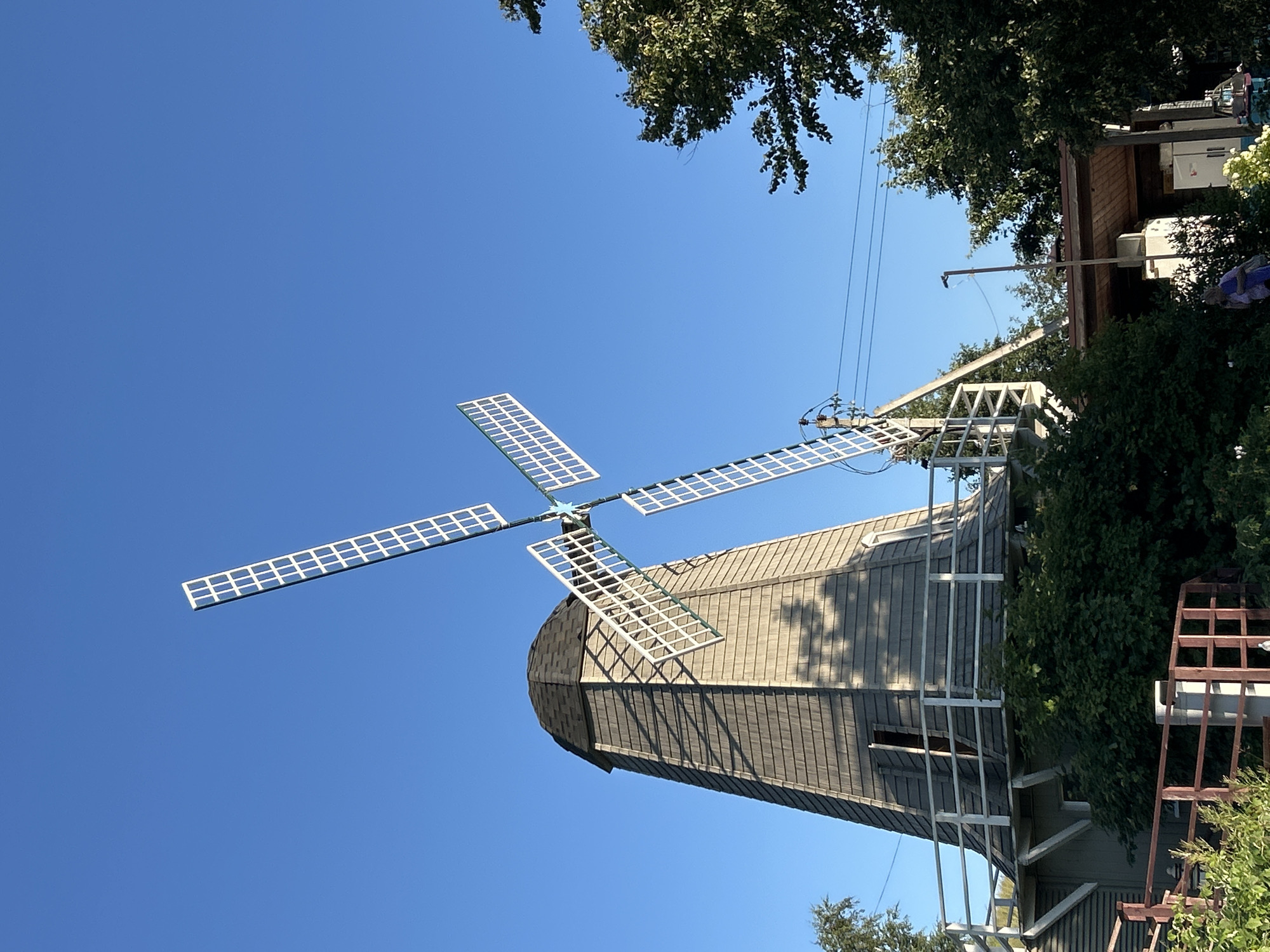 The image captures a traditional Dutch windmill standing against a clear blue sky. The windmill's tower is clad in weathered grey wooden shingles, with a dark grey, shingled conical roof. A white railing encircles a platform partway up the tower, which has an open doorway. The windmill's four large, white-framed sails are partially visible, angled against the sky. Lush green foliage and trees surround the base of the windmill. In the foreground, a red wooden fence and more greenery are visible. To the right, a covered outdoor area with a wooden roof and supporting pillars is present, with a white refrigerator and some decorative items visible. A person in a purple garment is partially visible in the lower right quadrant of the image, near the covered area. The scene suggests a rural or semi-rural setting, possibly a historical site or a farm. The image is taken on a clear day, with sunlight casting shadows on the windmill. The windmill is located in the village of Romashkovo, Russia.
