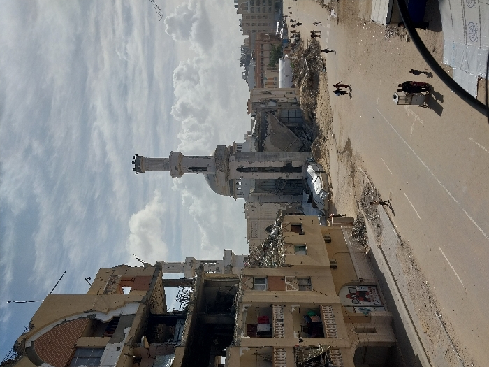 The image displays a street scene in Khan Yunis, Palestinian Territory, characterized by extensive structural destruction under a partially cloudy sky. In the foreground to the left, a multi-story building, possibly residential or commercial, exhibits significant damage, with large portions of its facade and internal structures collapsed, revealing debris-filled interiors. Clothes are visible hanging on balconies of this building, and a partially obscured sign, potentially depicting "SQUID GAME," is on its ground floor.
Prominently featured in the mid-ground is a mosque, also heavily damaged, with its tall, slender minaret largely intact at the top, where several birds are perched. The mosque's main structure, including its dome, shows signs of considerable collapse and disarray. The street below is paved but largely covered in dirt and scattered debris, though white road markings are discernible. Piles of rubble and remnants of destroyed structures line the street and extend into the background, where more damaged buildings are visible.
Several individuals are present on the street and sidewalks: one person walks alone on the left sidewalk, two individuals walk together in the center of the street, and a group consisting of three adults and a child are gathered around a small, white vendor's cart on the right side of the road. Further down the street, multiple other individuals are seen walking. A flock of birds is flying in the upper right portion of the sky.