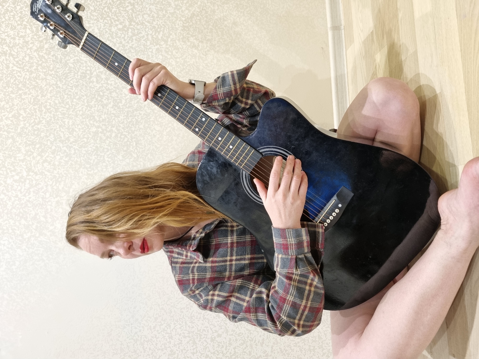 A fair-skinned person with light brown hair is seated on the floor and playing an acoustic guitar. The person is wearing a gray, red, and tan plaid long-sleeved shirt. The guitar is dark blue and black. The person is playing the guitar with their left hand on the neck and their right hand on the strings. The person is seated with their legs crossed, and the guitar is resting on their lap. The floor is light wood. The background is a light beige wall. It is not possible to confirm the location of Chaikovsky, Russia from the image provided.